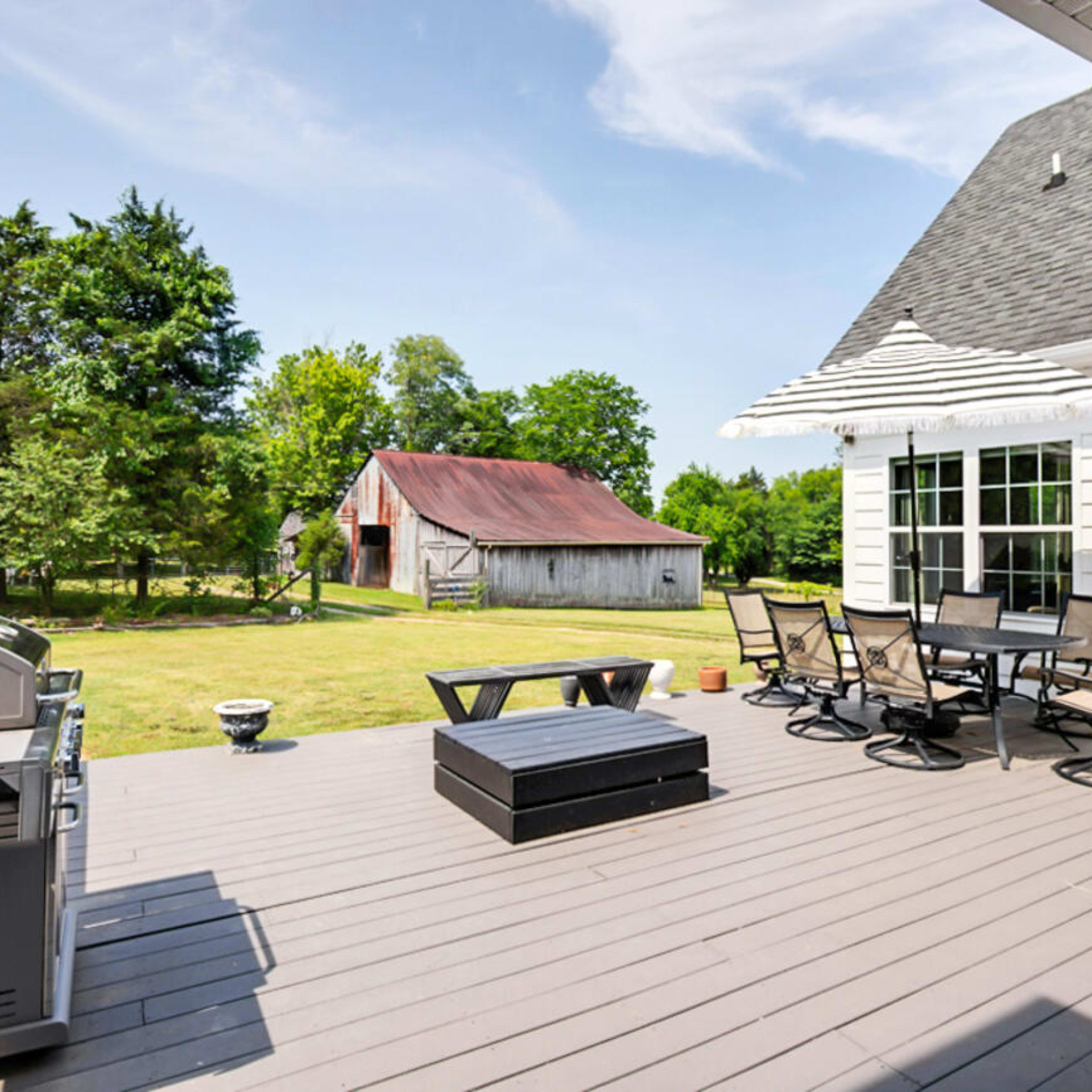 A spacious deck features a grill, patio furniture, and a view of a green lawn with a barn in the background.