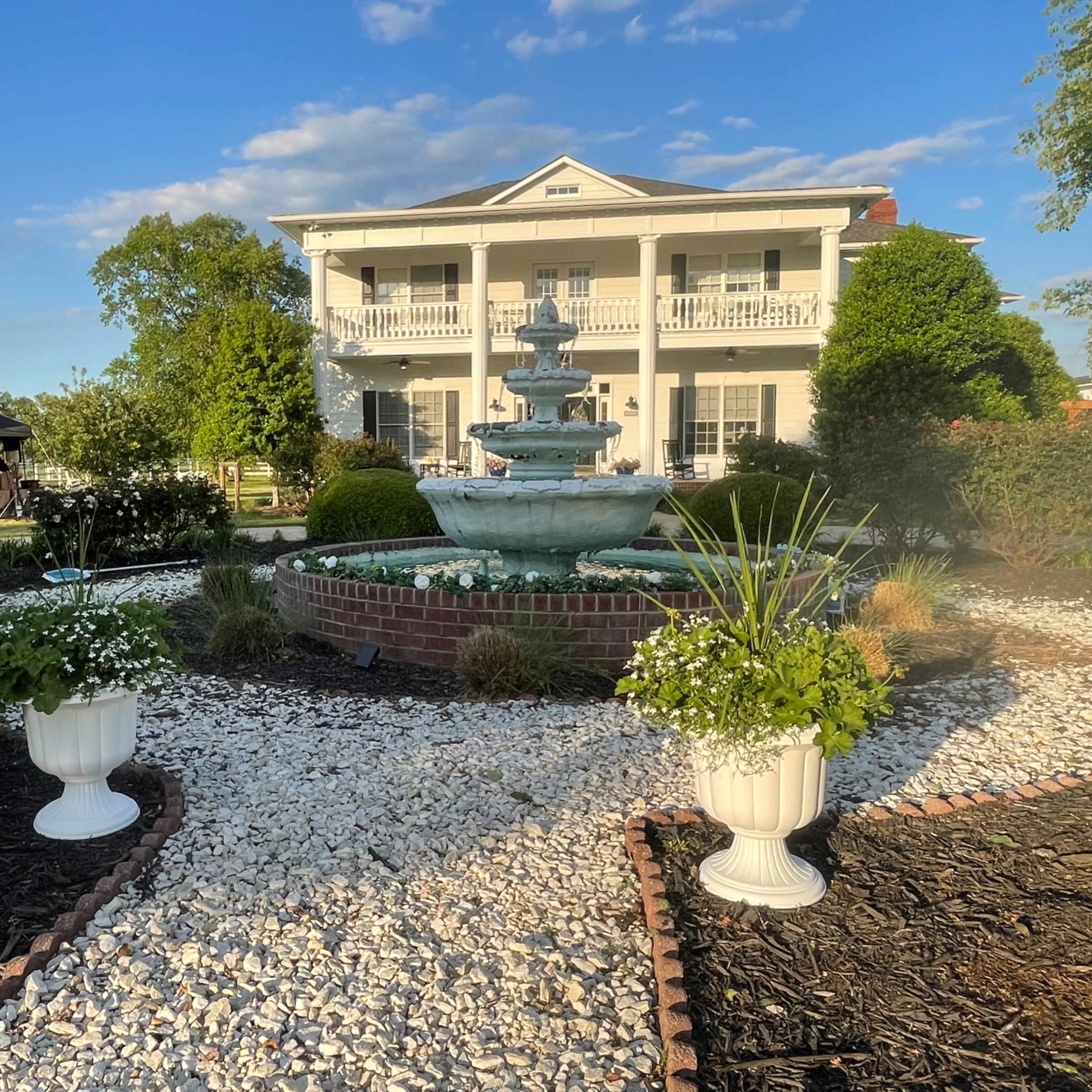 A multi-tiered fountain is surrounded by a landscaped garden featuring potted plants and a large colonial-style house in the background.