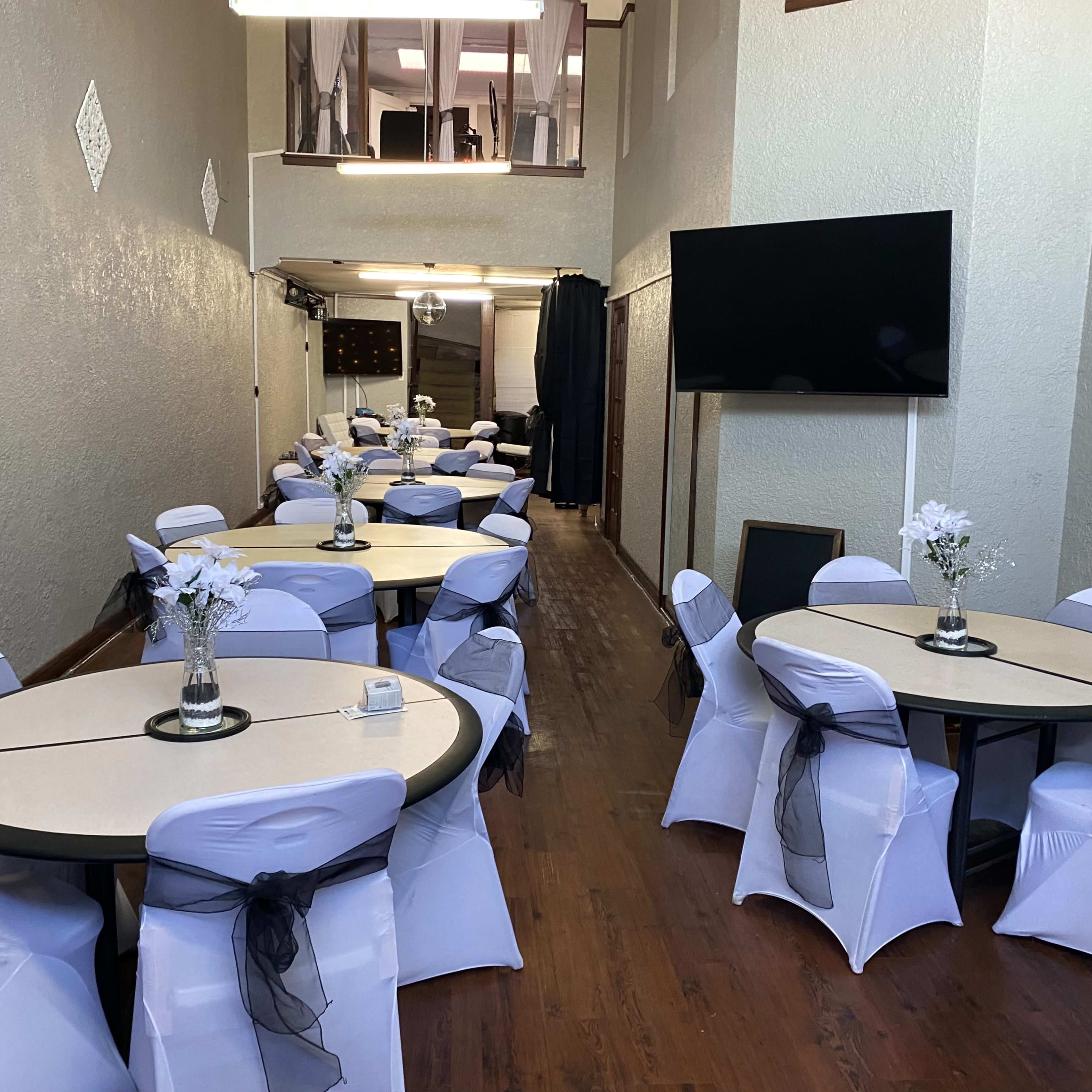 A decorated event space with round tables covered in white tablecloths and black sashes, arranged neatly along the length of the room.
