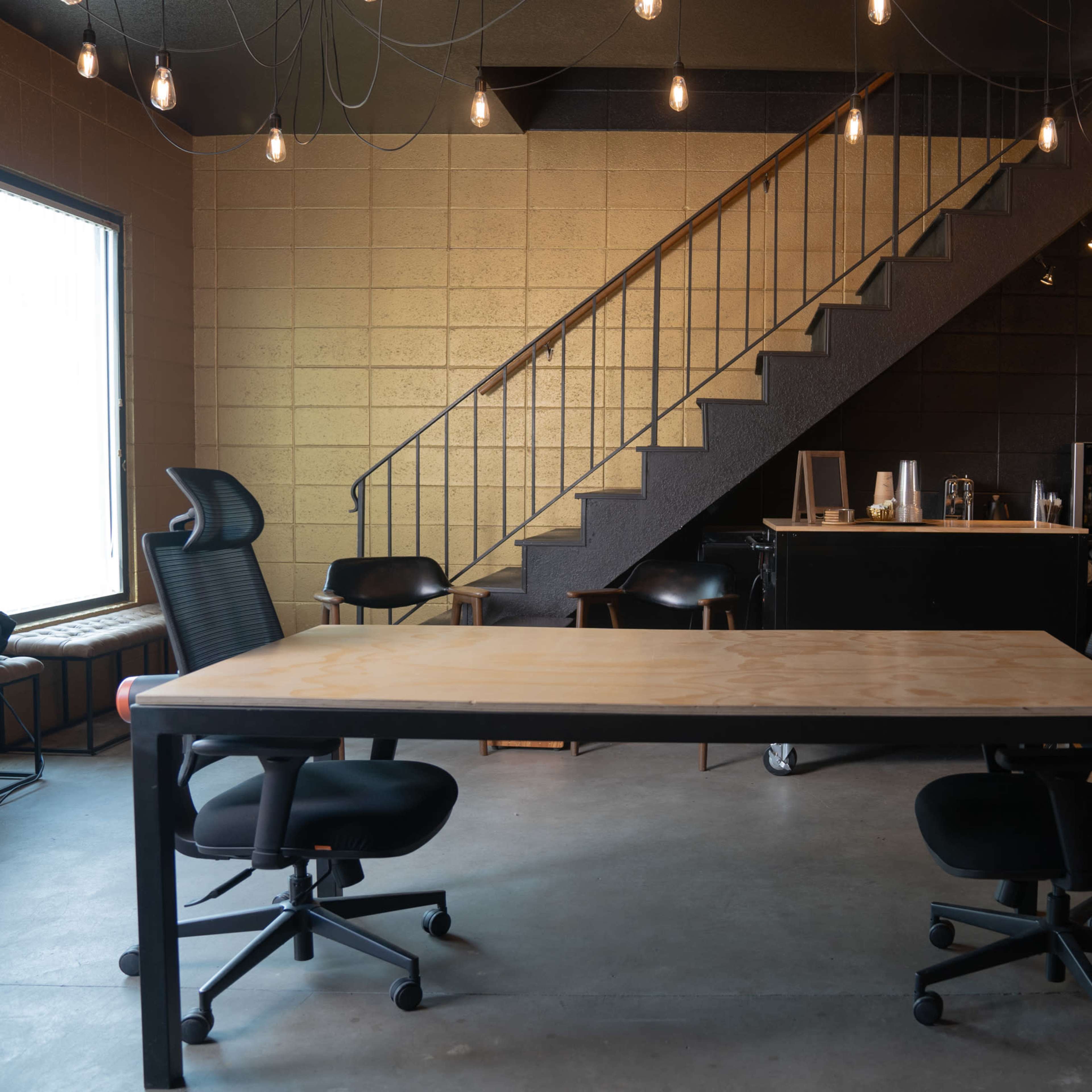 An office space features a large table with two chairs, a staircase in the background, and bar seating with various beverages nearby.