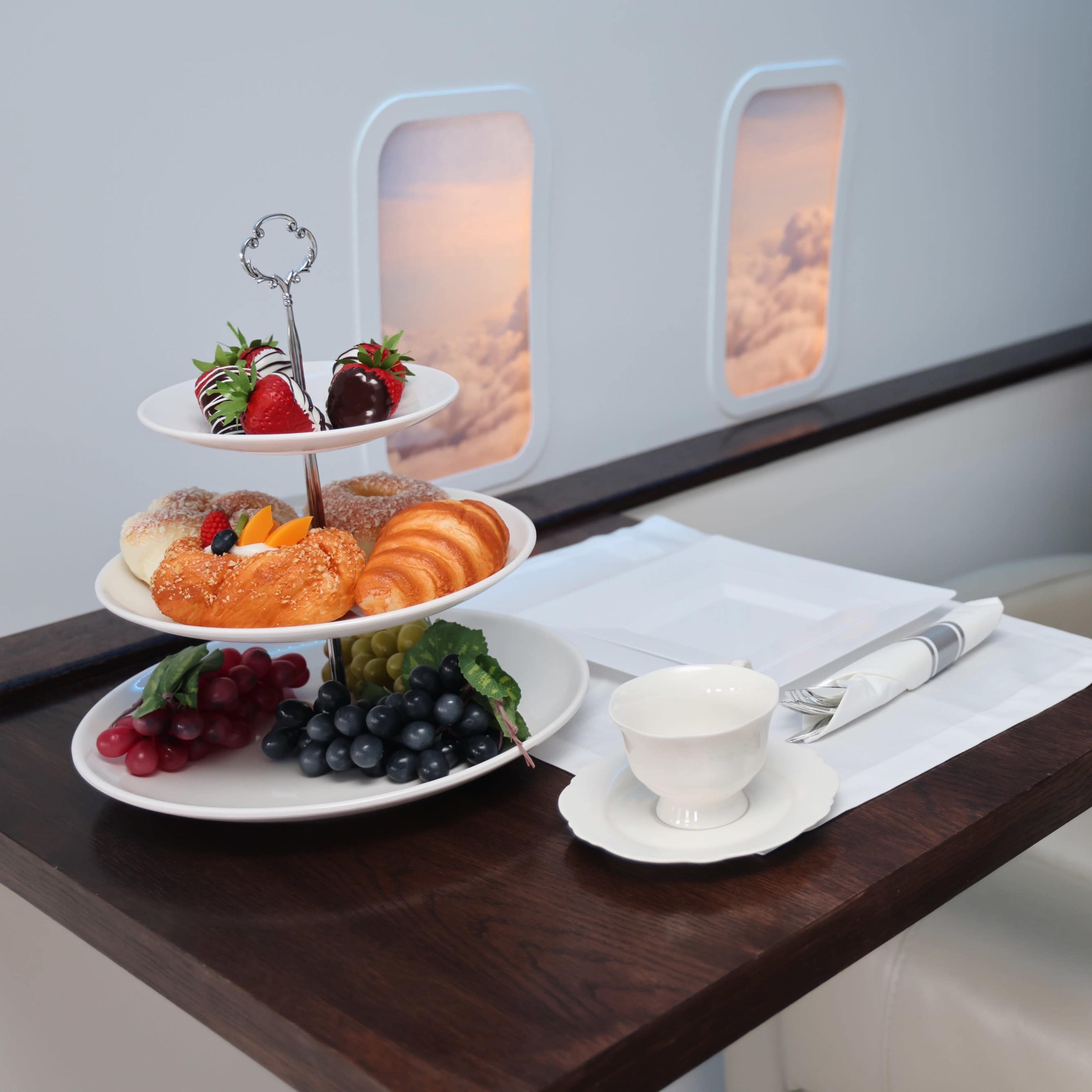 A tiered stand with pastries, fruits, and berries is placed on a wooden table next to a white cup in an airplane seat beside windows showing clouds.