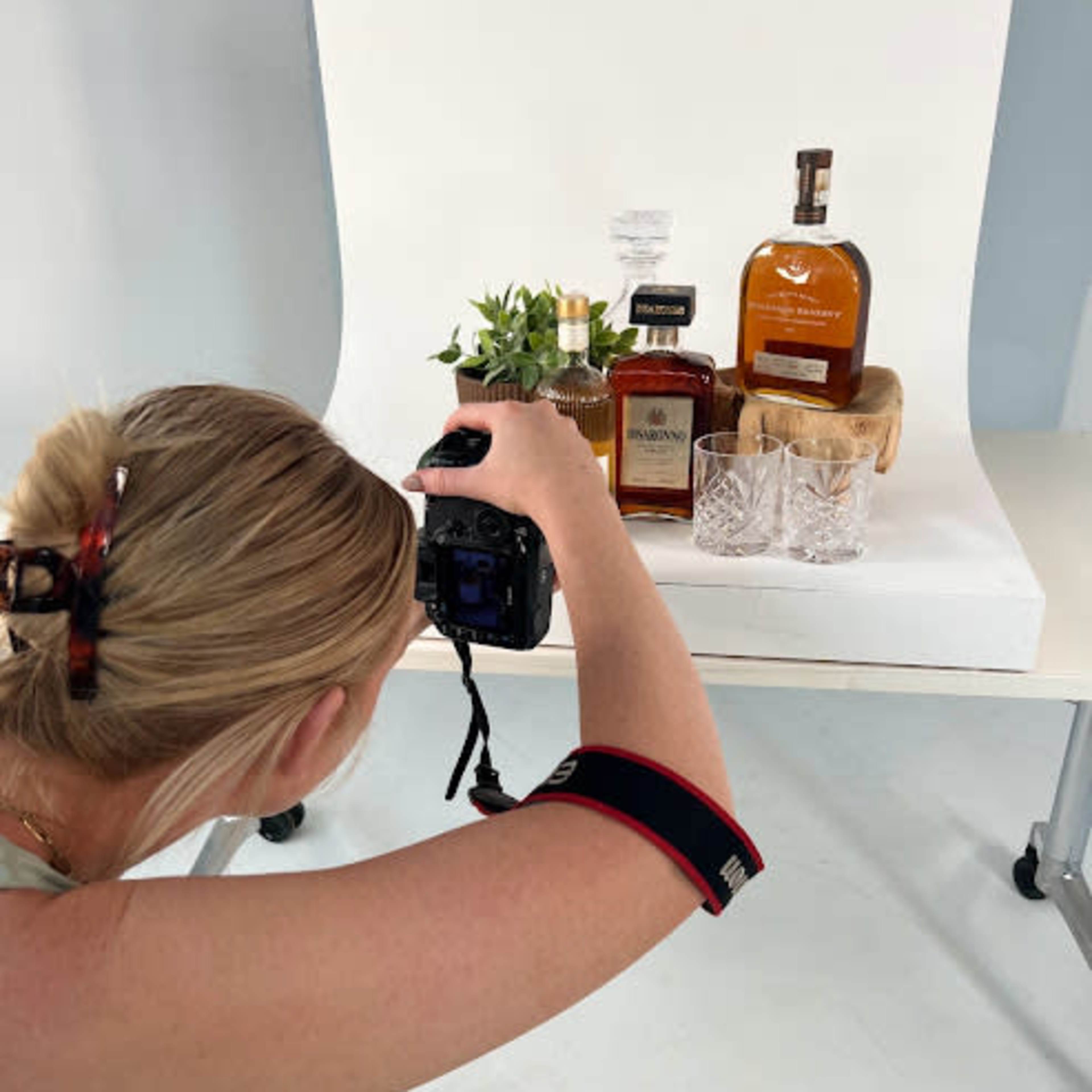 A person is taking a photograph of bottles of liquor and decorative items arranged on a white backdrop.