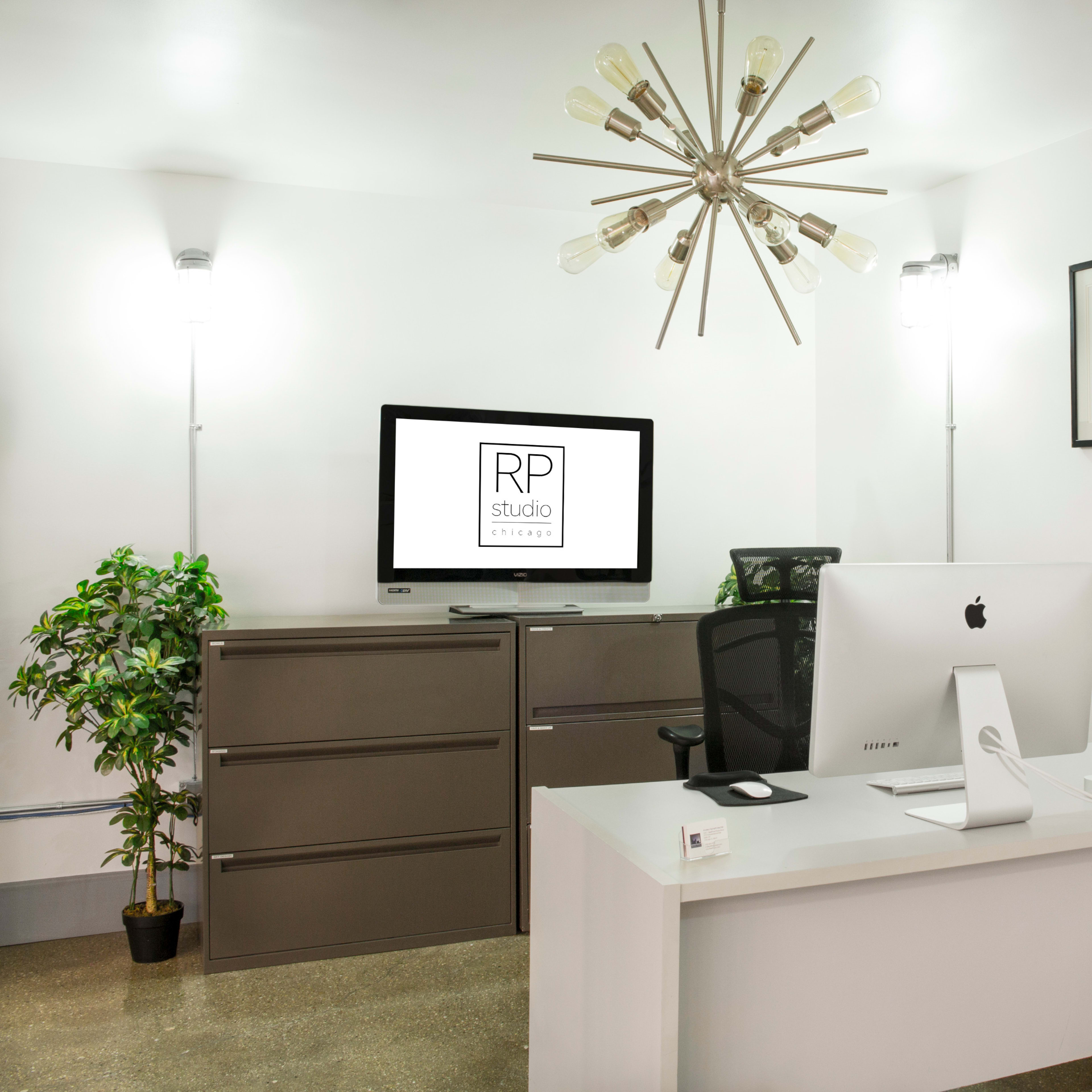 A modern office space featuring a desk with an Apple computer, a television monitor on a cabinet, a plant in the corner, and industrial-style lighting fixtures.