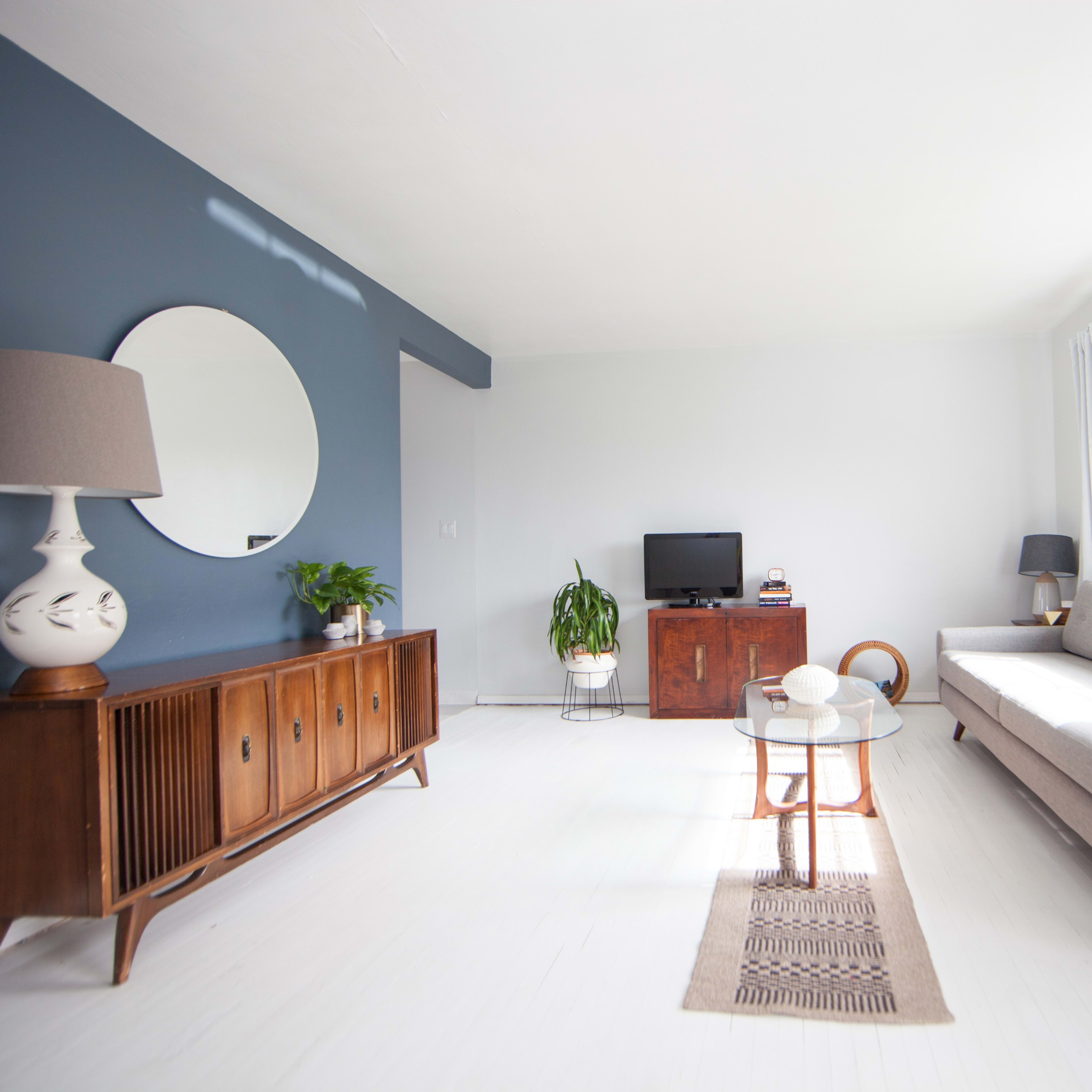 A modern living room with a gray sofa, a round mirror on a blue wall, a wooden sideboard, and a coffee table positioned in front of a window.
