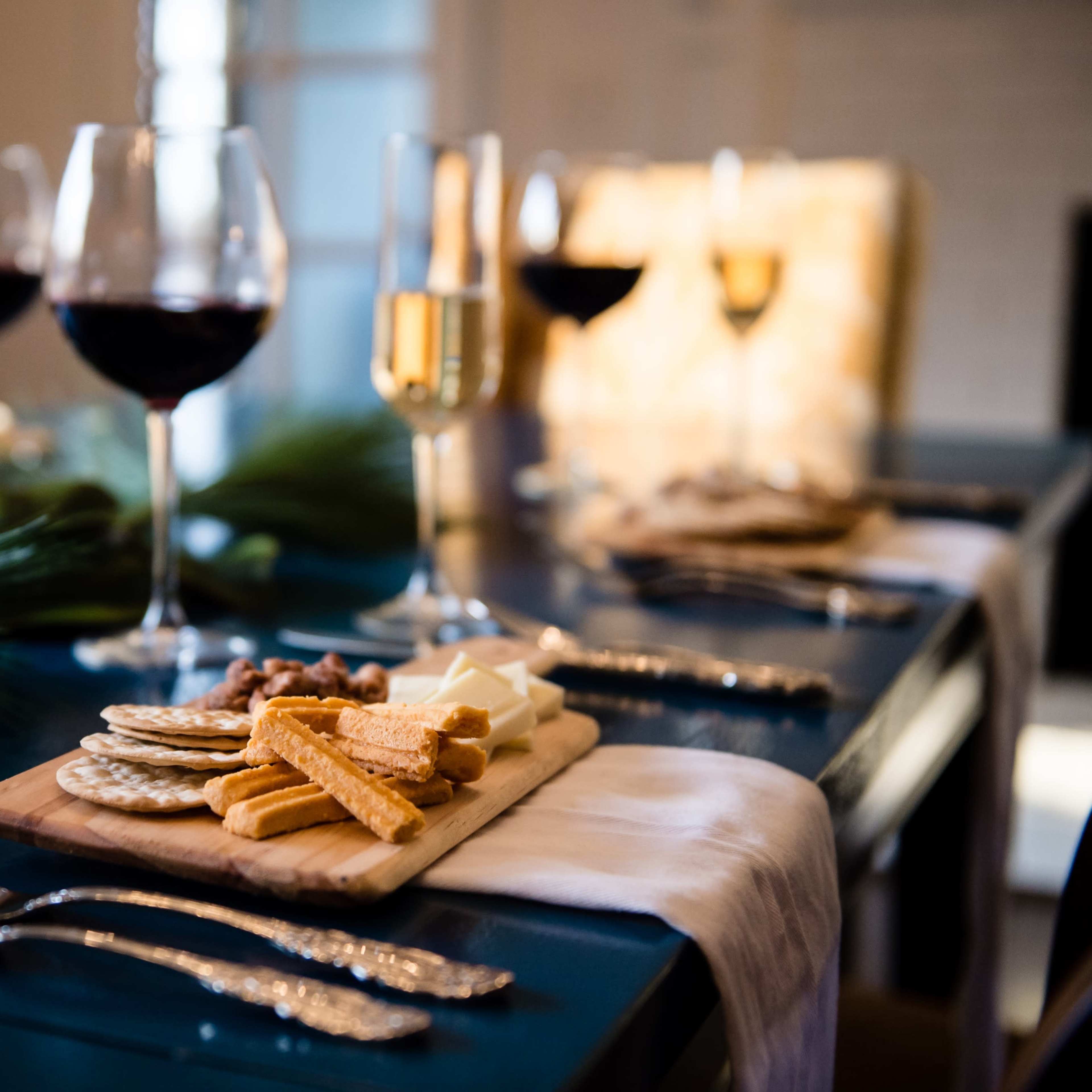 A wooden platter with an assortment of cheeses and crackers is set on a dining table adorned with wine glasses and greenery.