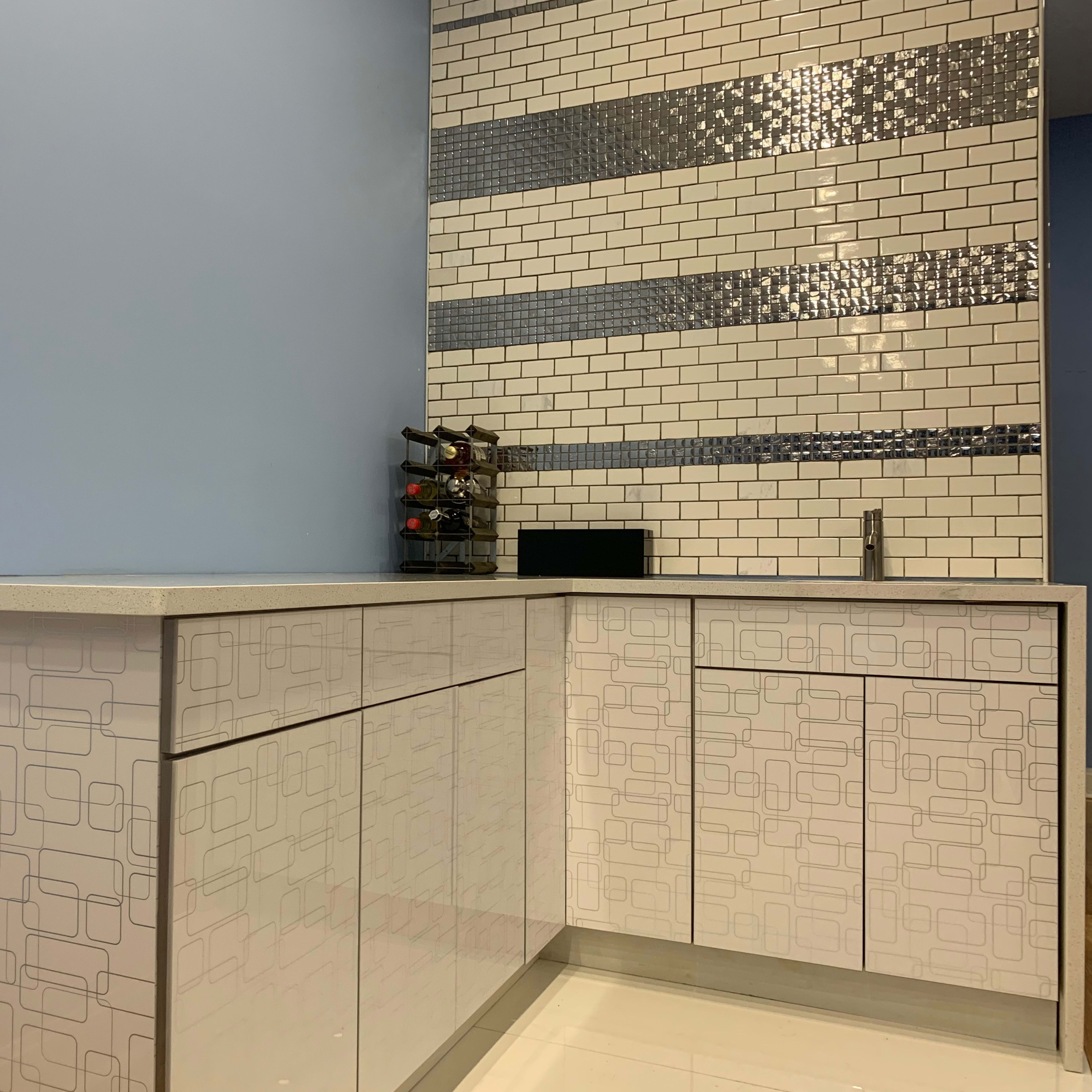 A modern kitchen corner featuring a countertop with patterned cabinets and a tiled backsplash with alternating rows of white and metallic tiles.
