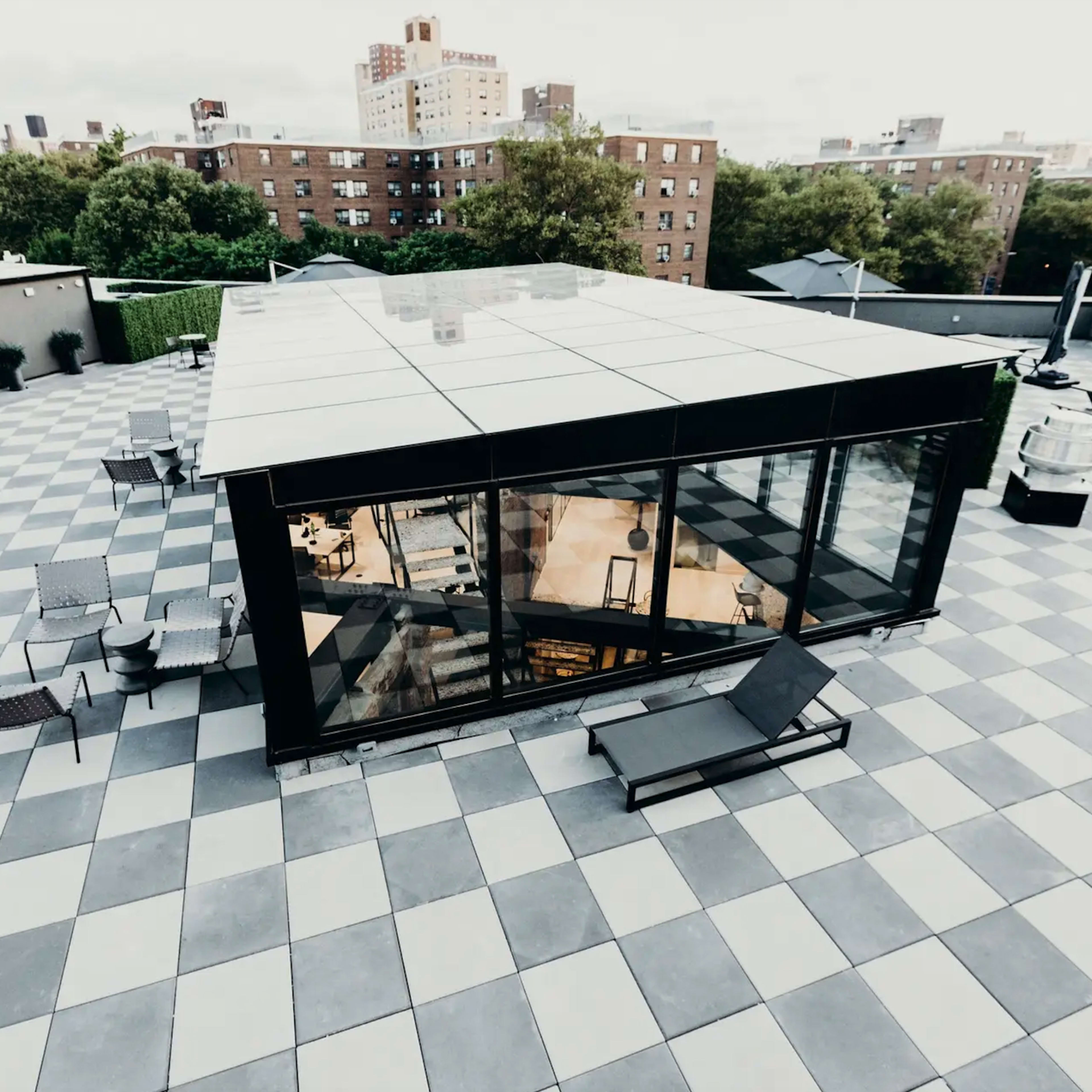 A modern glass pavilion sits atop a rooftop covered in gray and white tiled squares, surrounded by seating areas and city buildings in the background.
