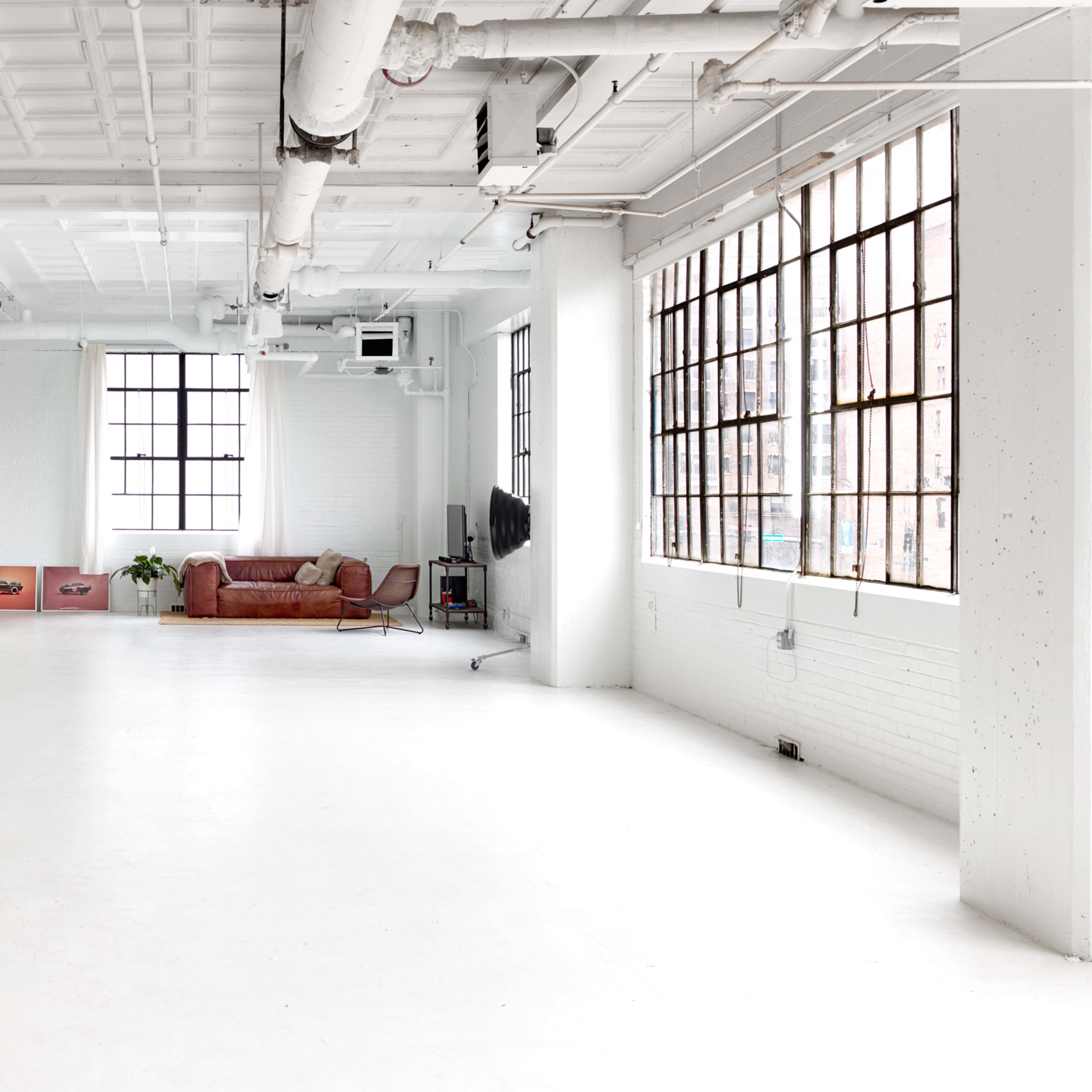 A spacious, minimalist industrial loft with large windows, white walls, and wooden floors, featuring a red couch in the background.