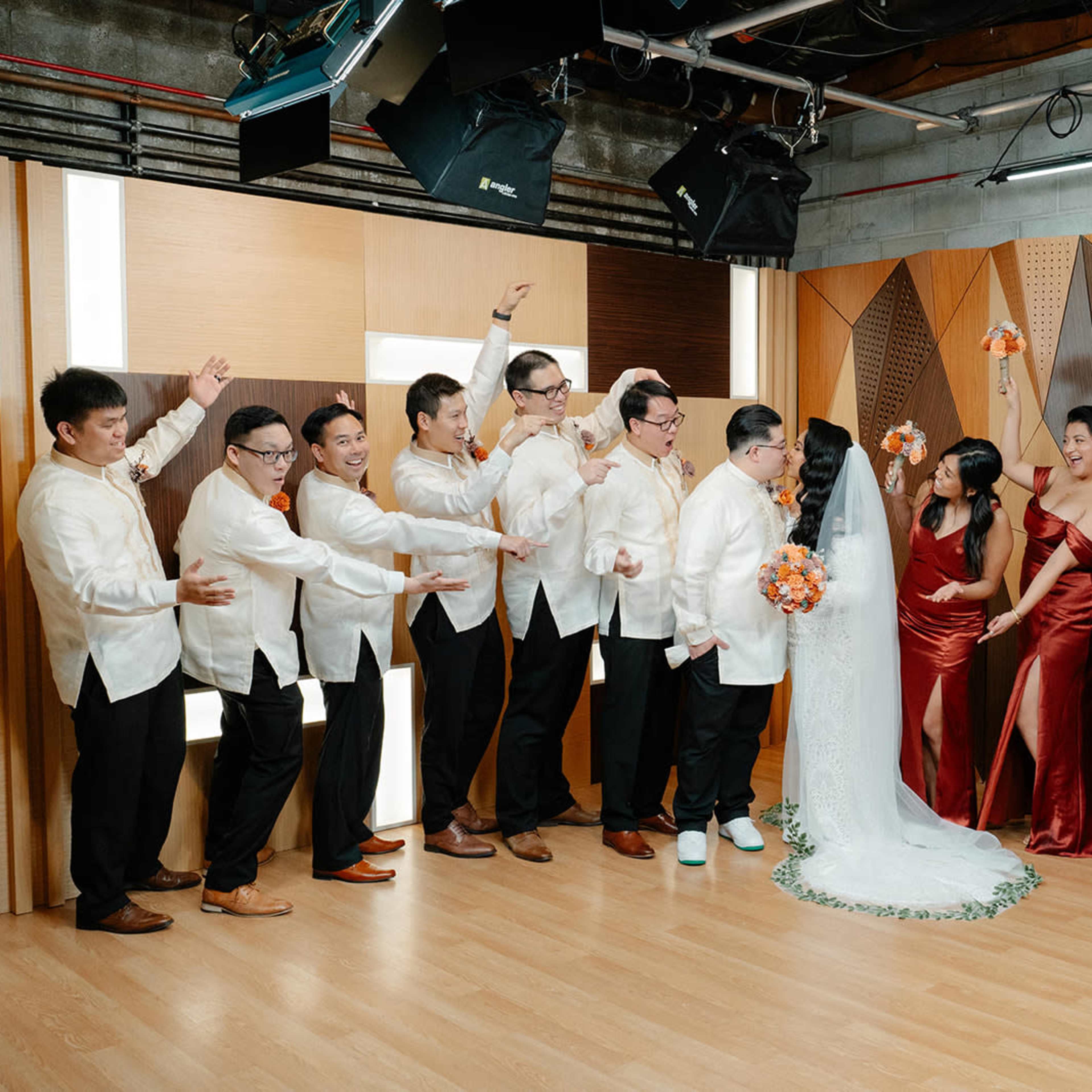 A wedding party poses together in a studio, with the bride and groom at the center surrounded by guests in formal attire.