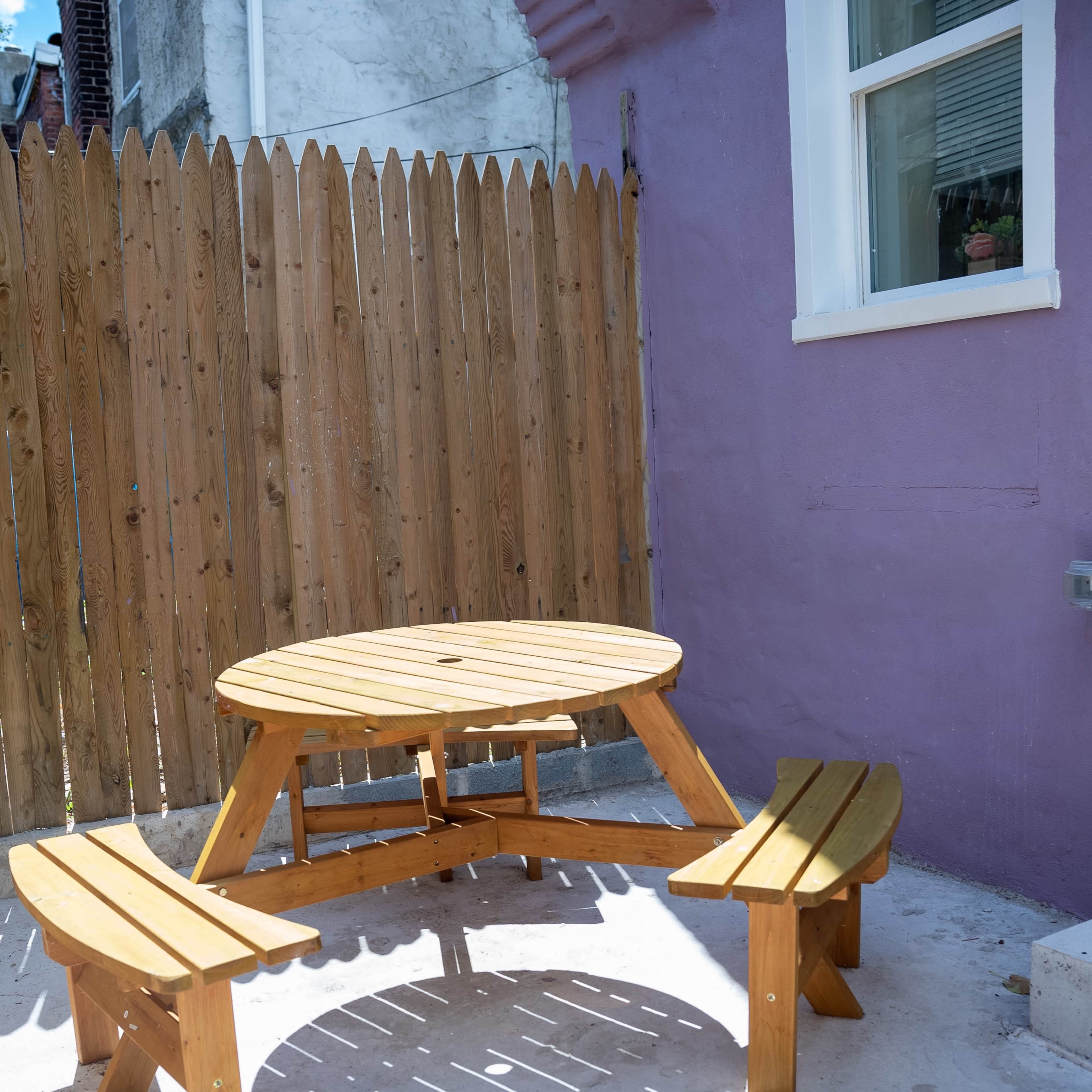 A round wooden picnic table with two attached benches is placed on a concrete surface beside a purple wall and a wooden fence.