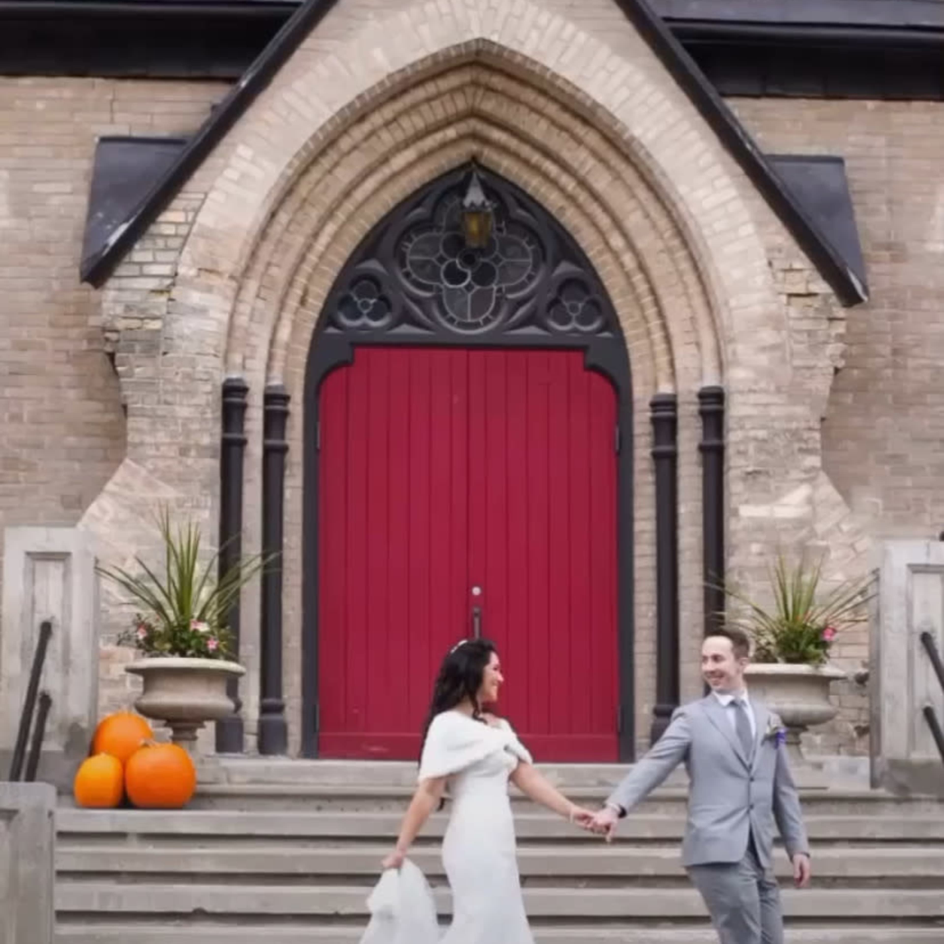 A bride and groom walk hand in hand down the steps of a cathedral, flanked by orange pumpkins.