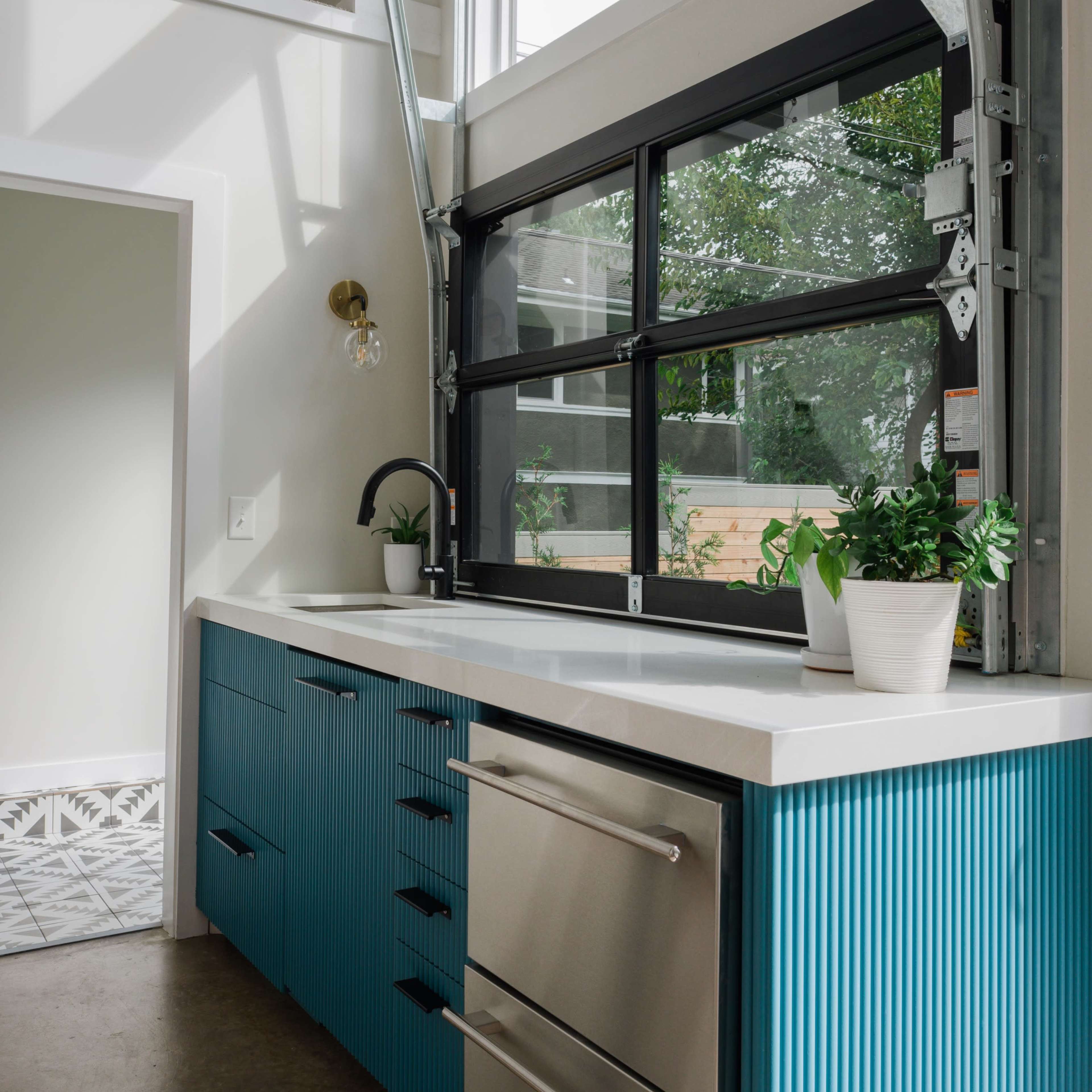 A modern kitchen features blue ribbed cabinetry, stainless steel appliances, and large windows that allow natural light to enter.