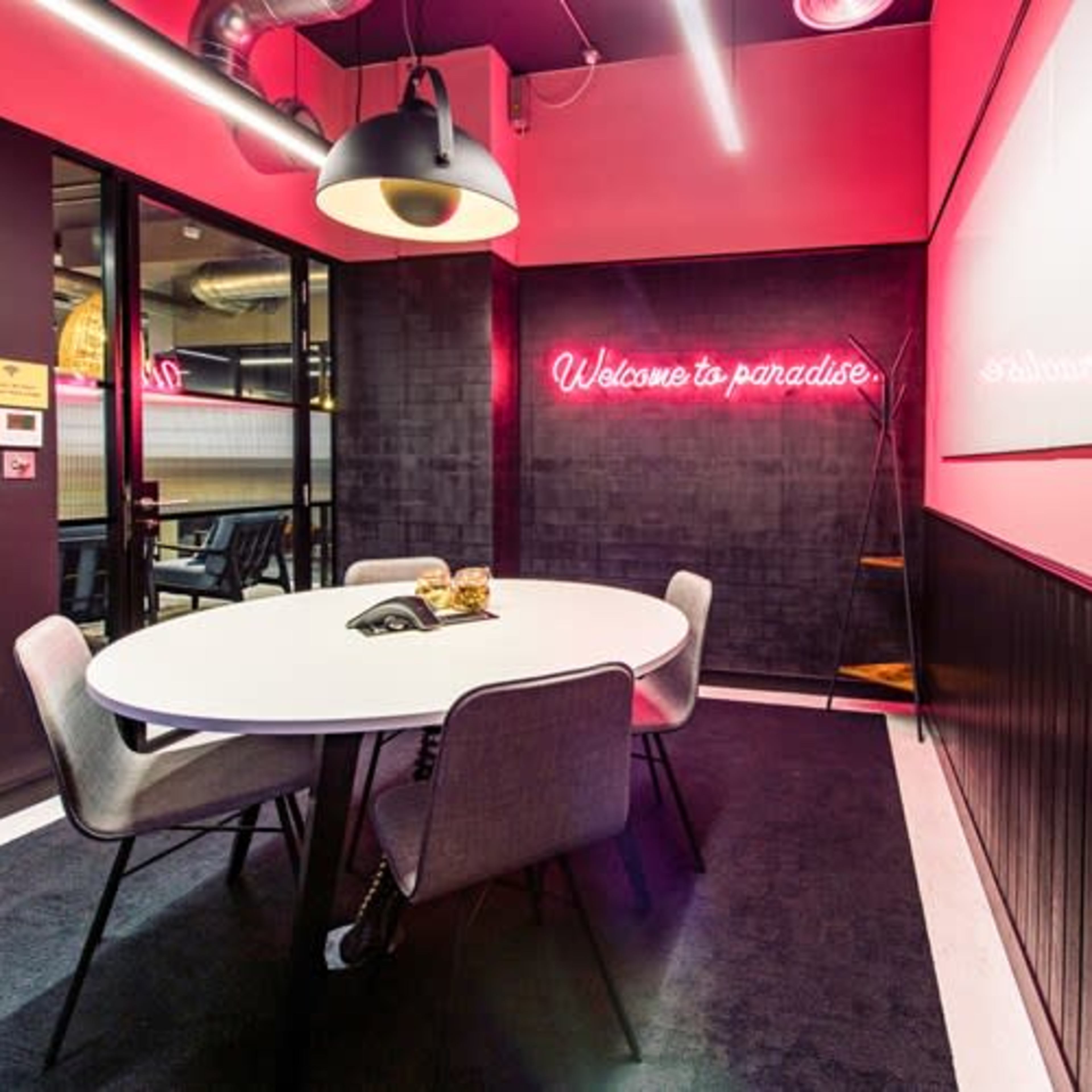 A modern meeting room features a round white table surrounded by upholstered chairs, with a neon sign reading "Welcome to paradise" on the wall.