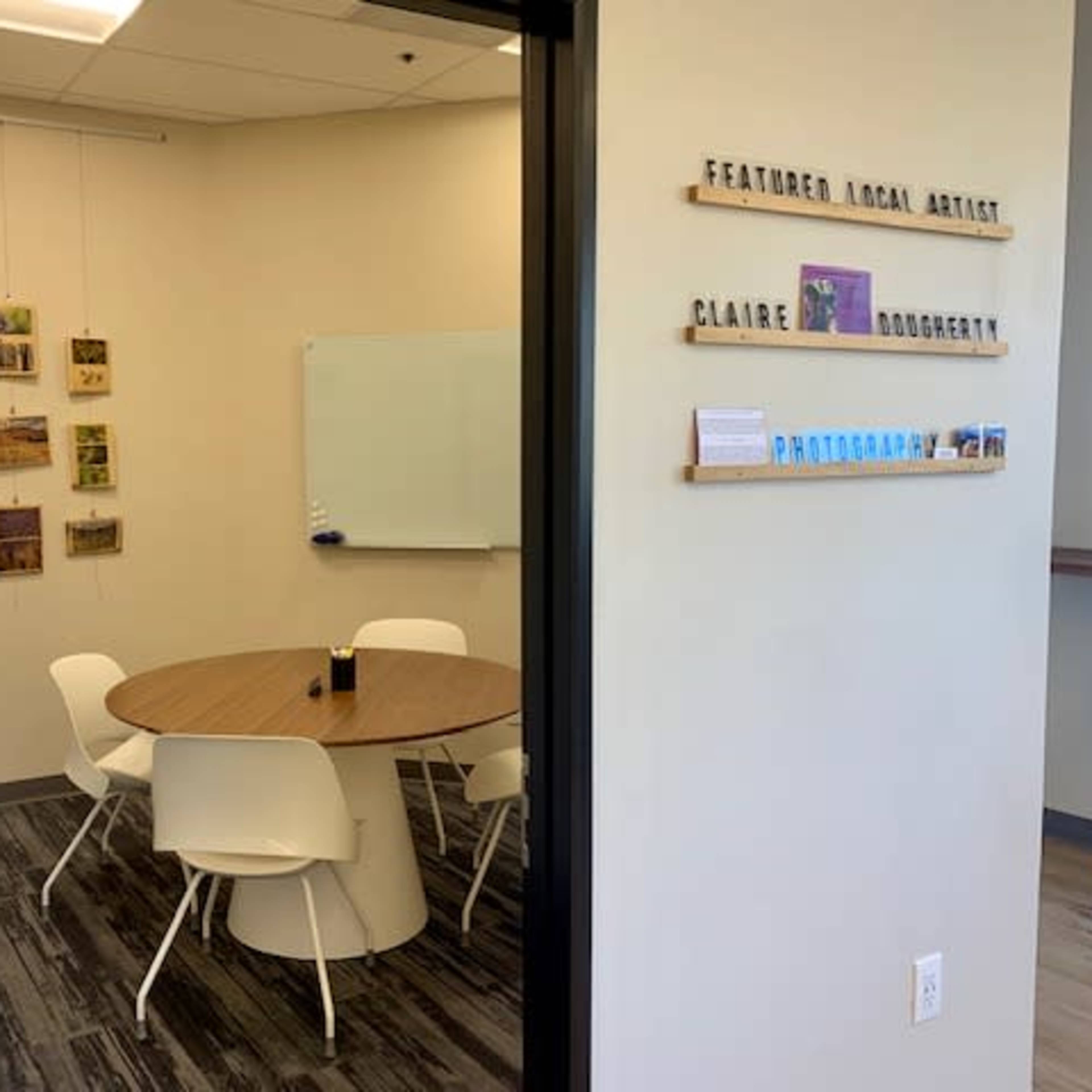 A small meeting room with a round wooden table surrounded by white chairs, featuring a gallery of photographs on the wall and a whiteboard.