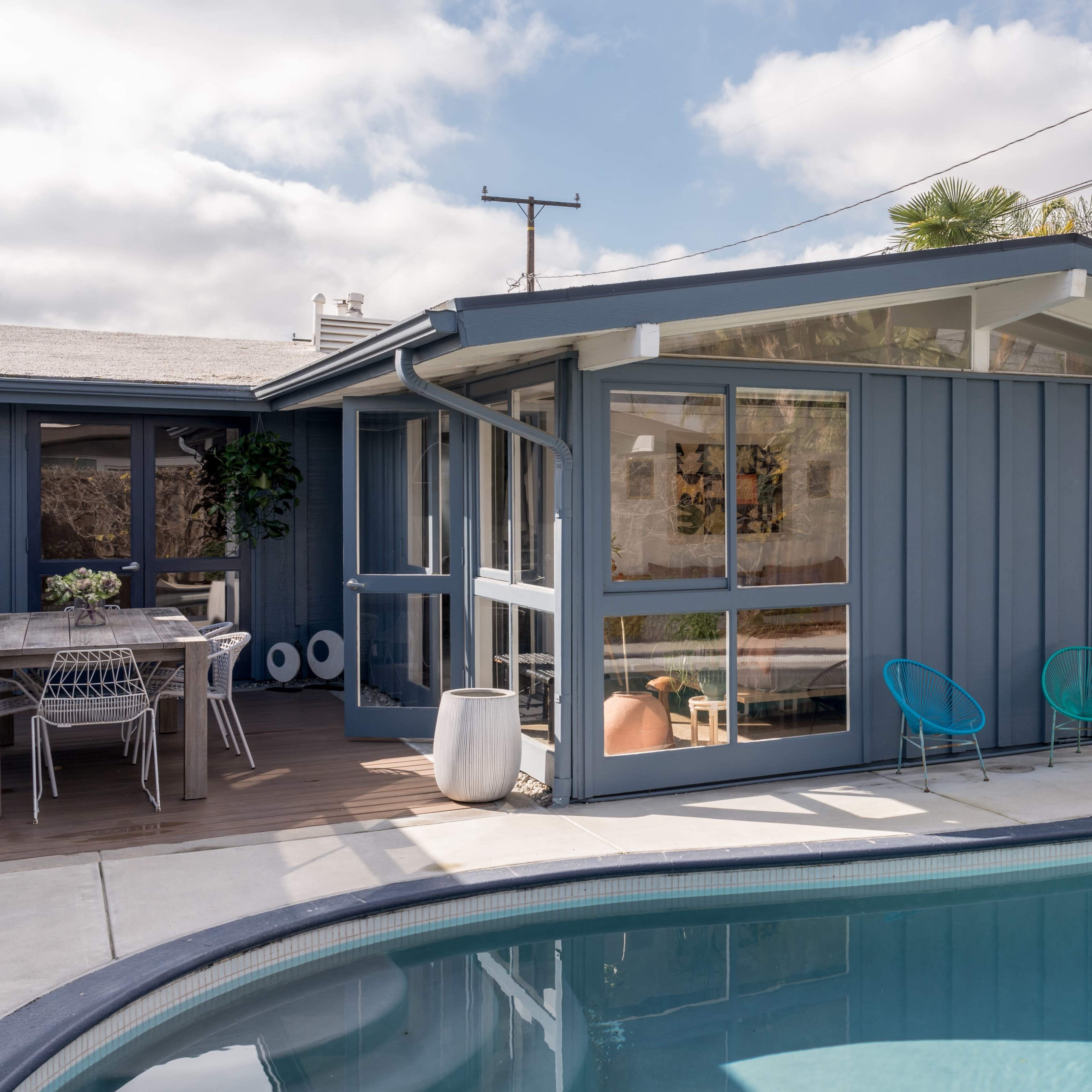 A modern, blue house with large windows overlooks a swimming pool surrounded by palm trees and outdoor seating.
