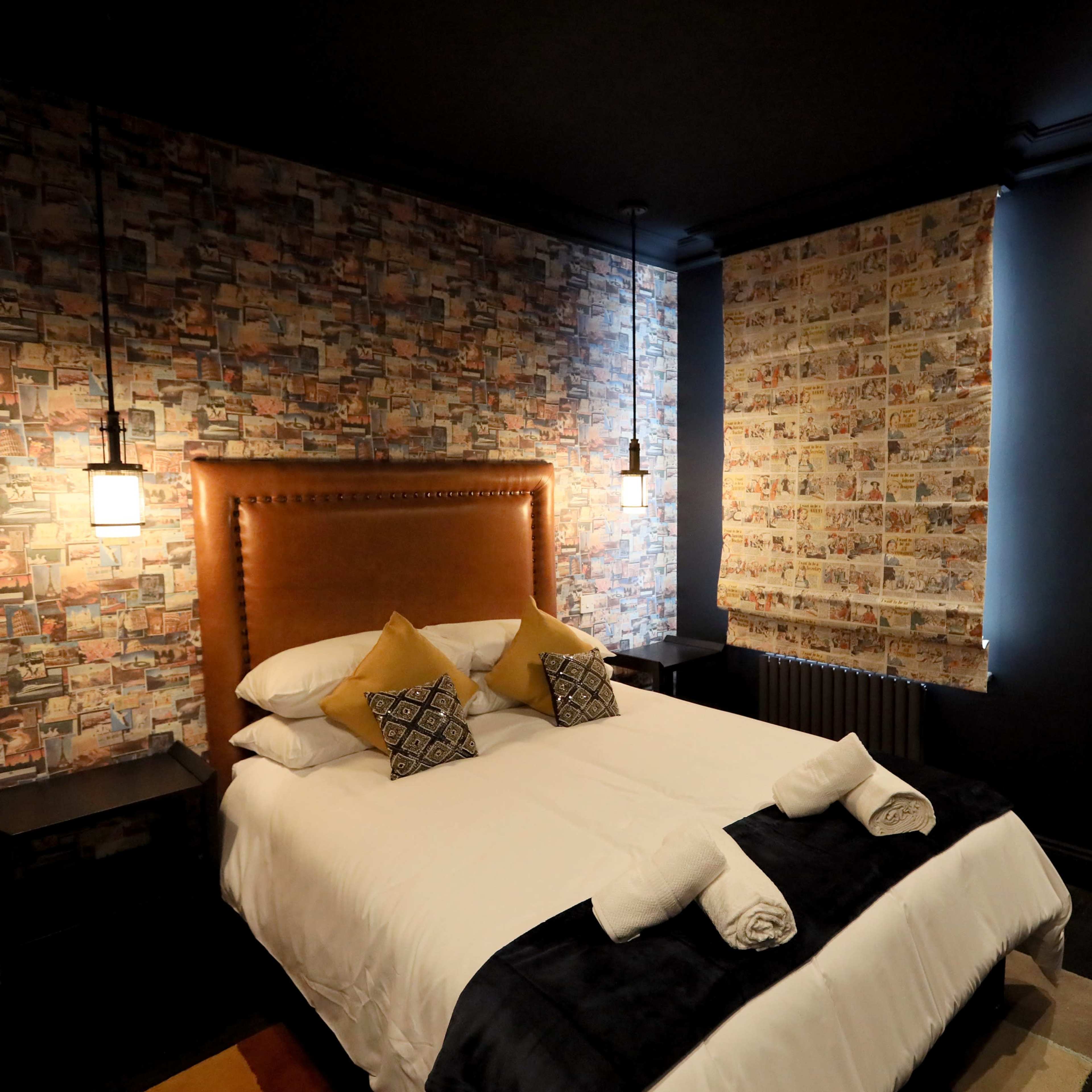 A stylish bedroom featuring a brown upholstered headboard, neatly made white bed with rolled towels, patterned wallpaper, and ambient lighting.
