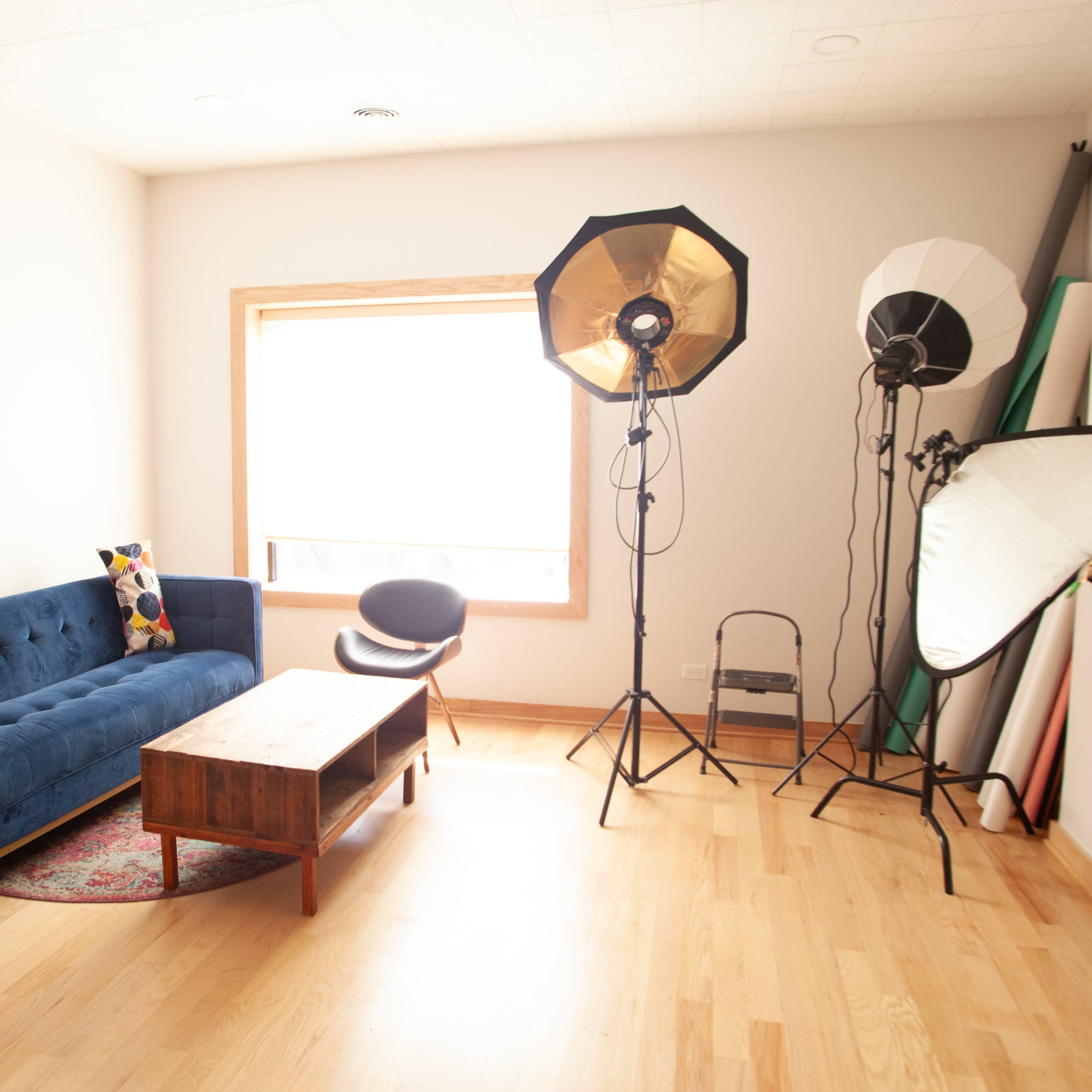 A cozy living room features a blue sofa, a wooden coffee table, and studio lighting equipment next to a large window.