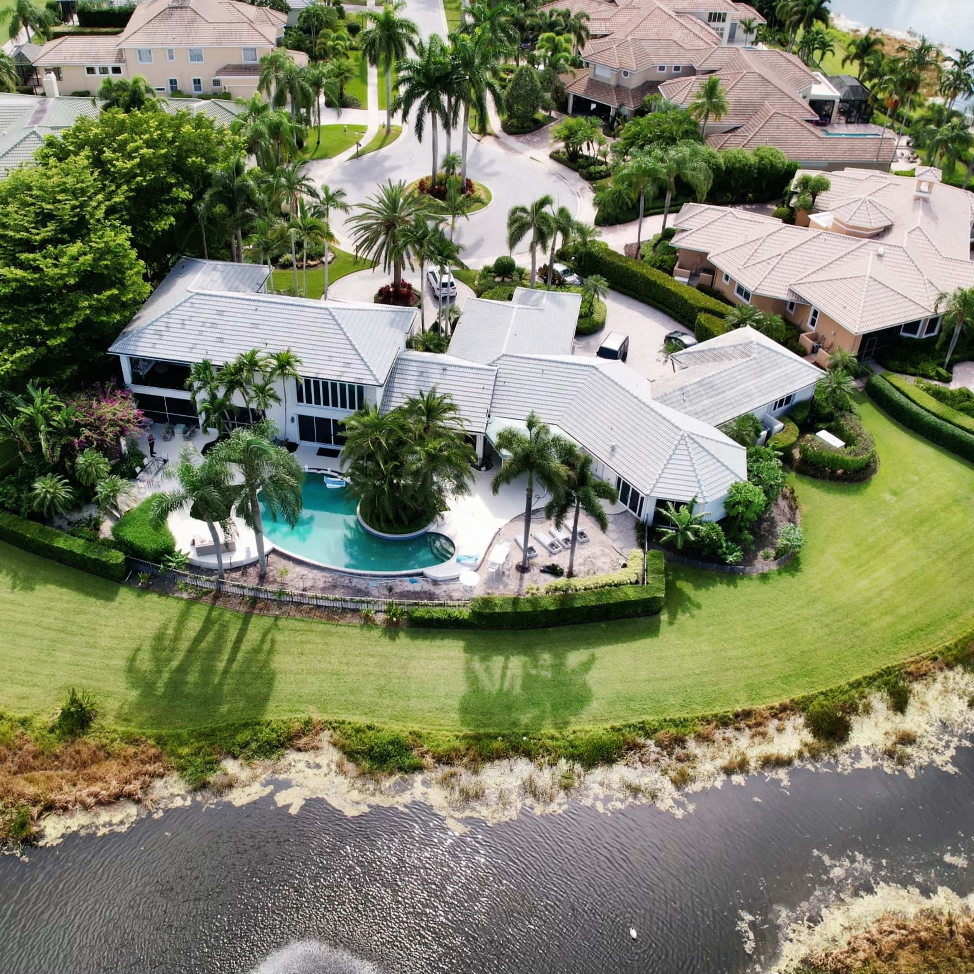 Aerial view of a waterfront property surrounded by a lush lawn and palm trees, with a pool and several nearby houses.