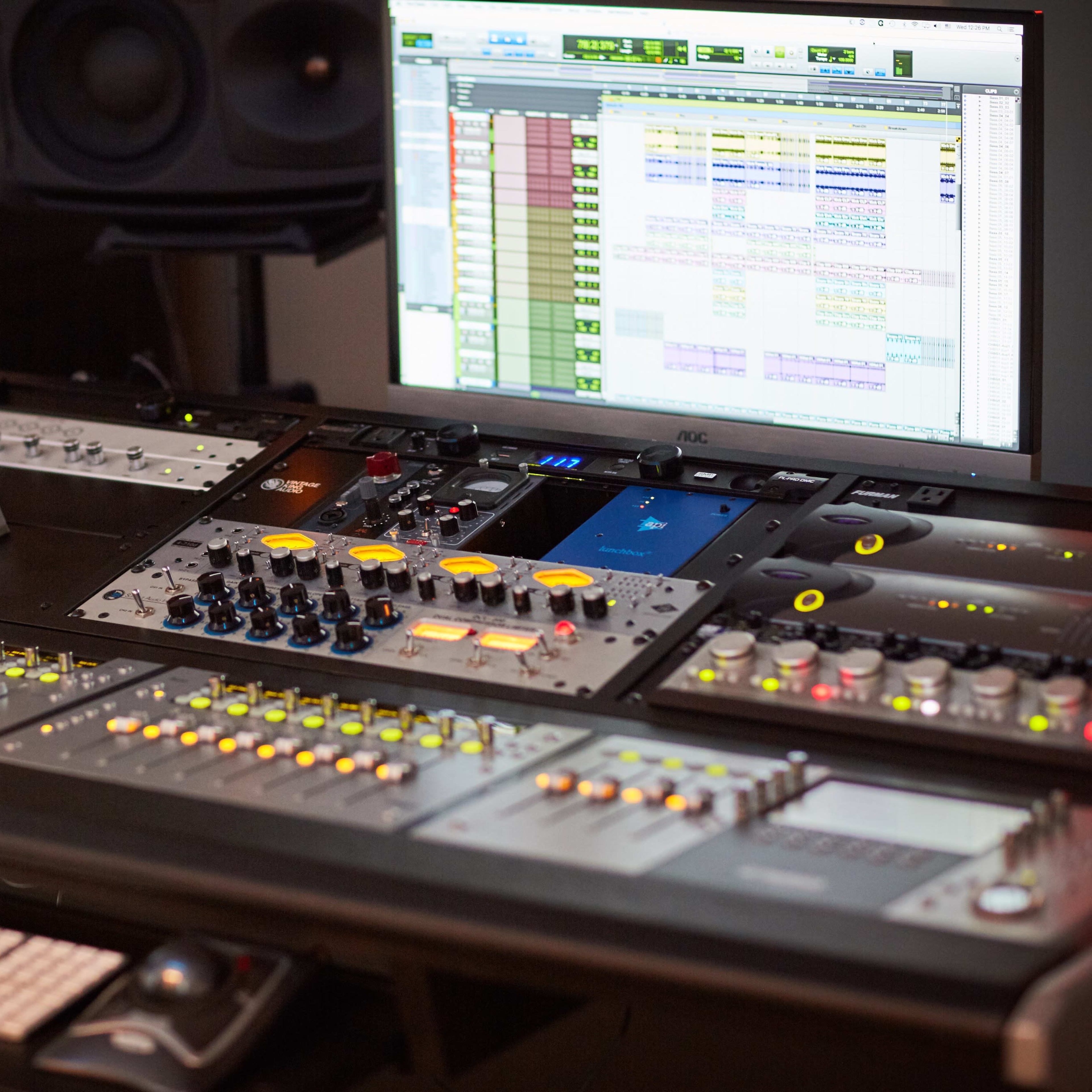 A recording studio workstation features a large mixing console, various audio control units, and a computer monitor displaying a digital audio workstation interface.