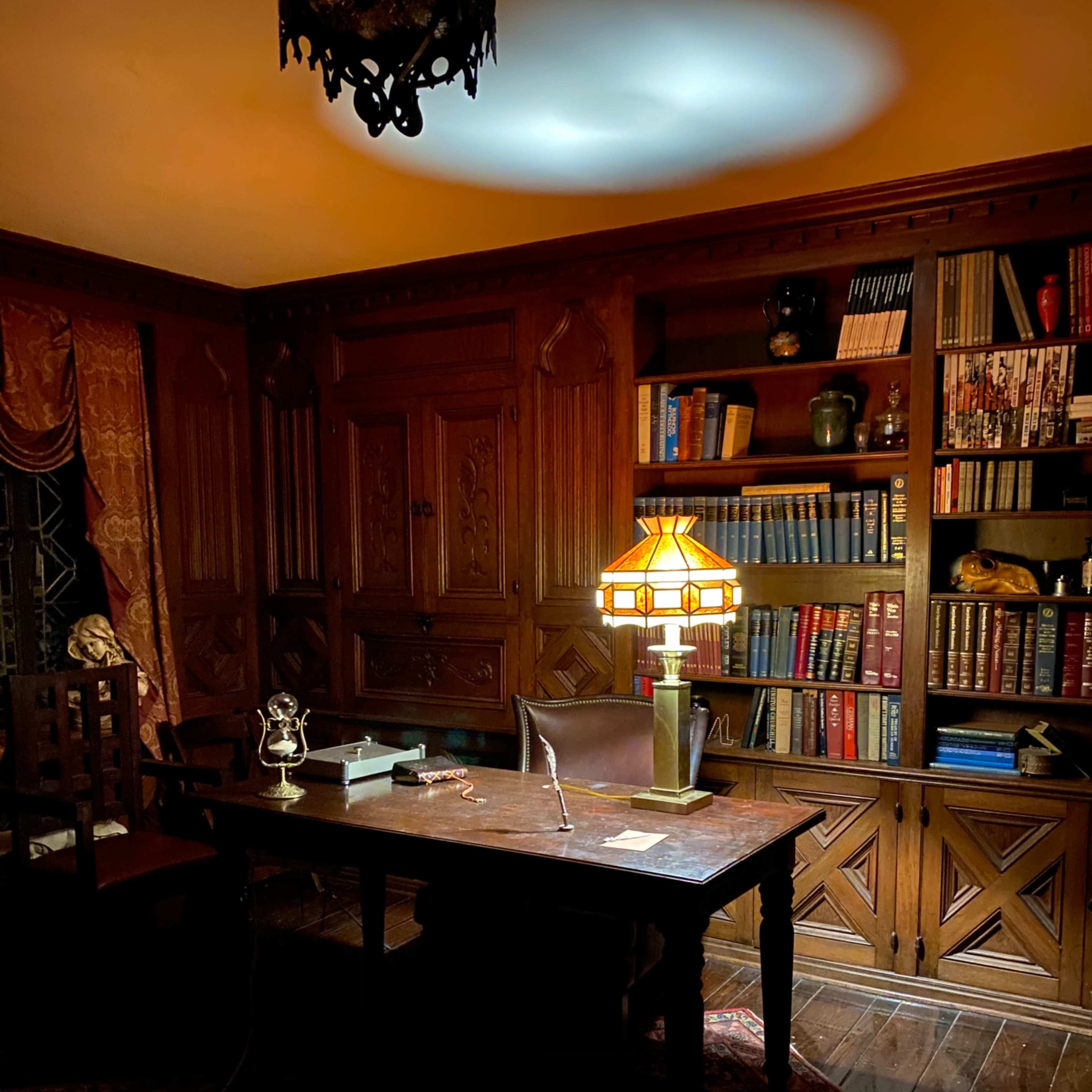 A dimly lit wooden office with a large desk, a classic lamp, and a bookshelf filled with various books.