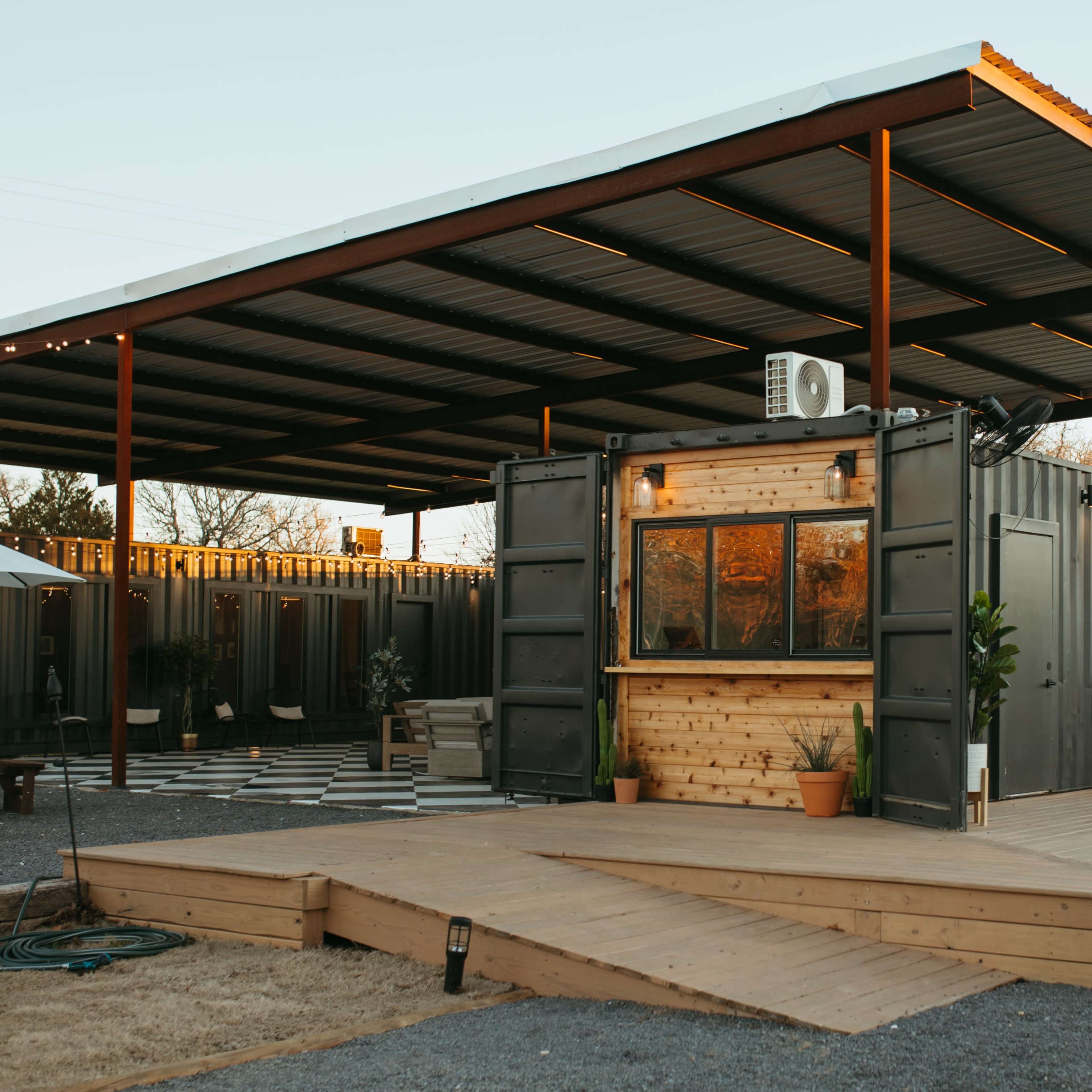 A modern, open-air structure featuring a wooden deck and a container-style building with large windows and outdoor seating.
