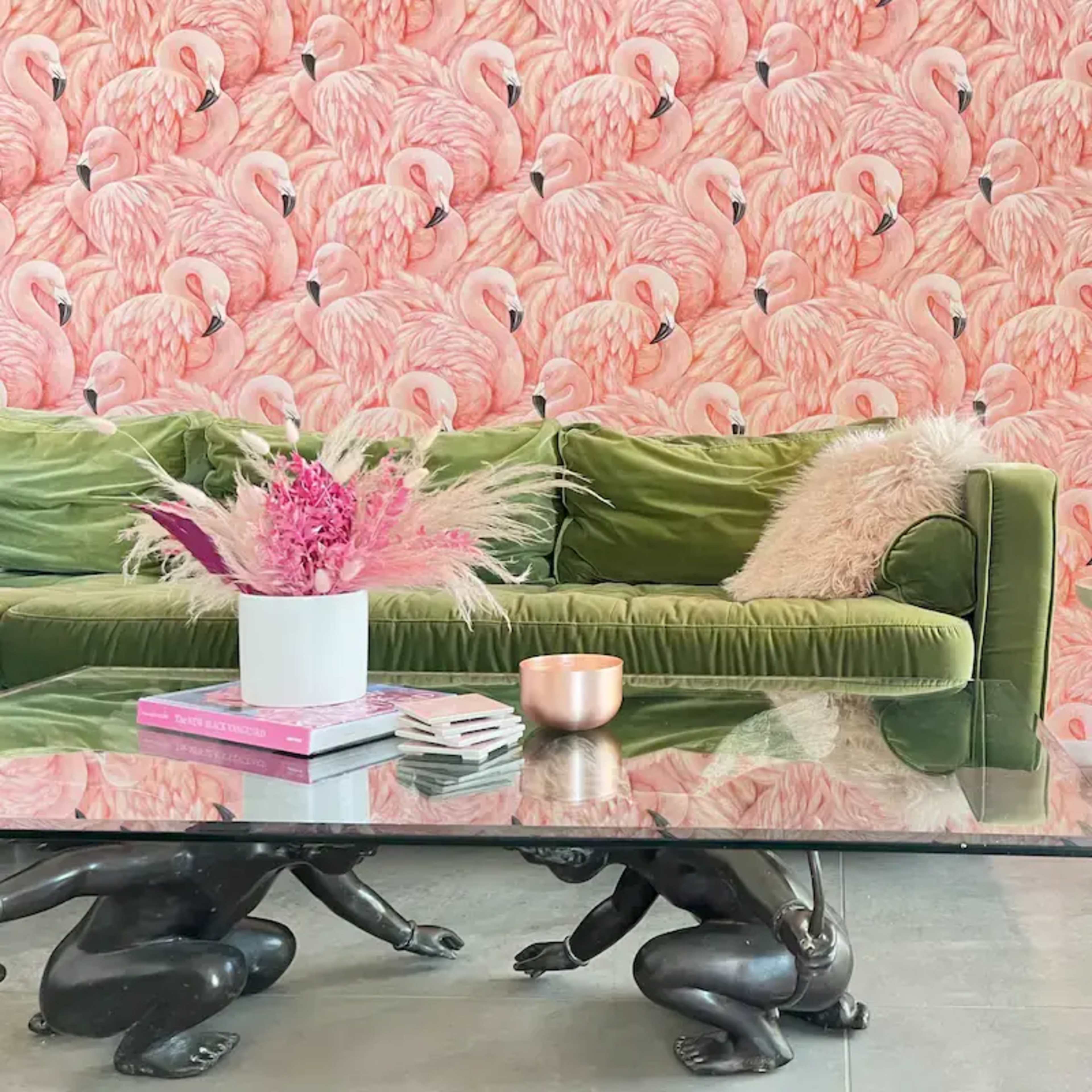 A green velvet sofa is positioned in front of a pink flamingo-patterned wallpaper, complemented by a glass coffee table and a rattan chair with a decorative cushion.