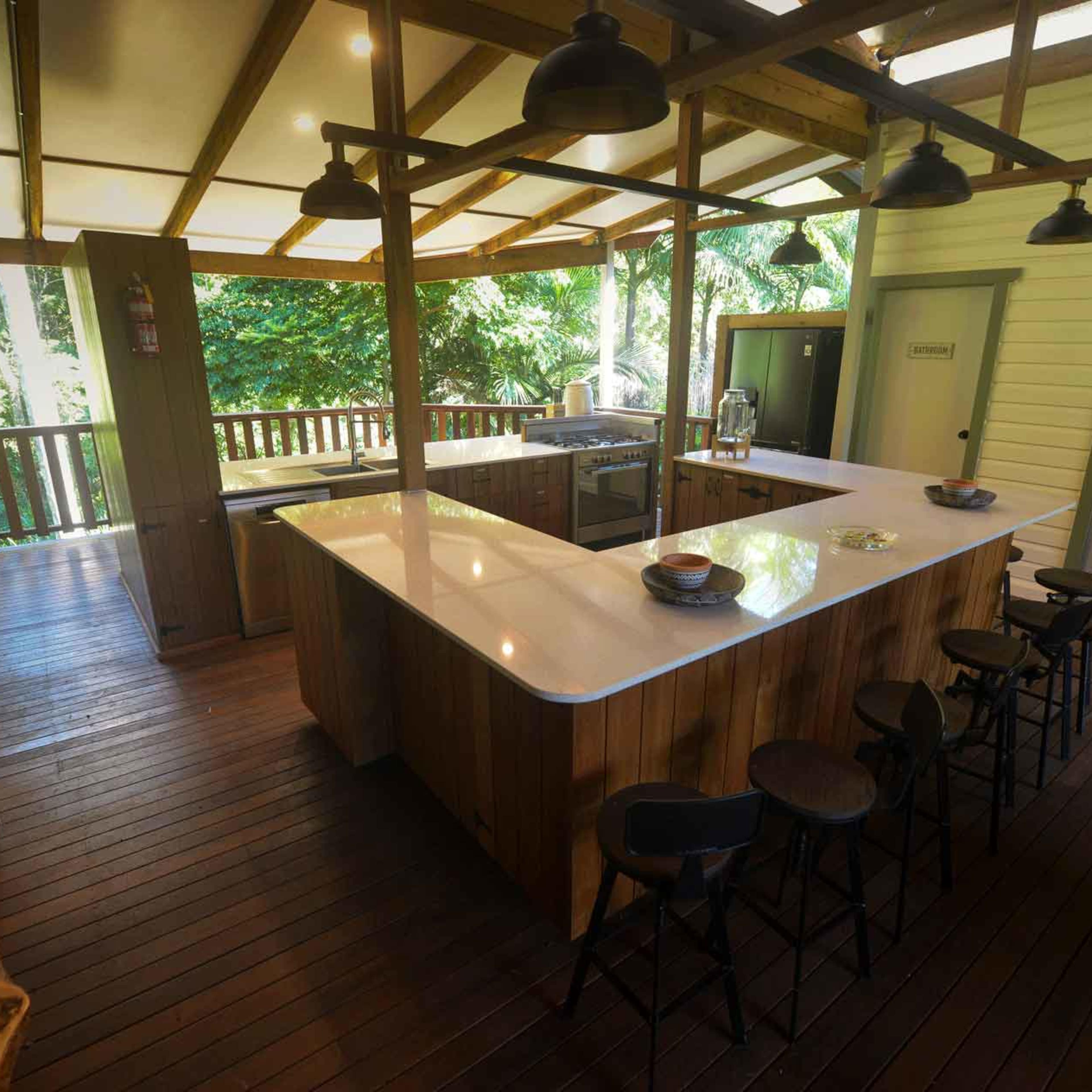 A modern kitchen with a large island and several bar stools, surrounded by wooden flooring and large windows overlooking greenery.