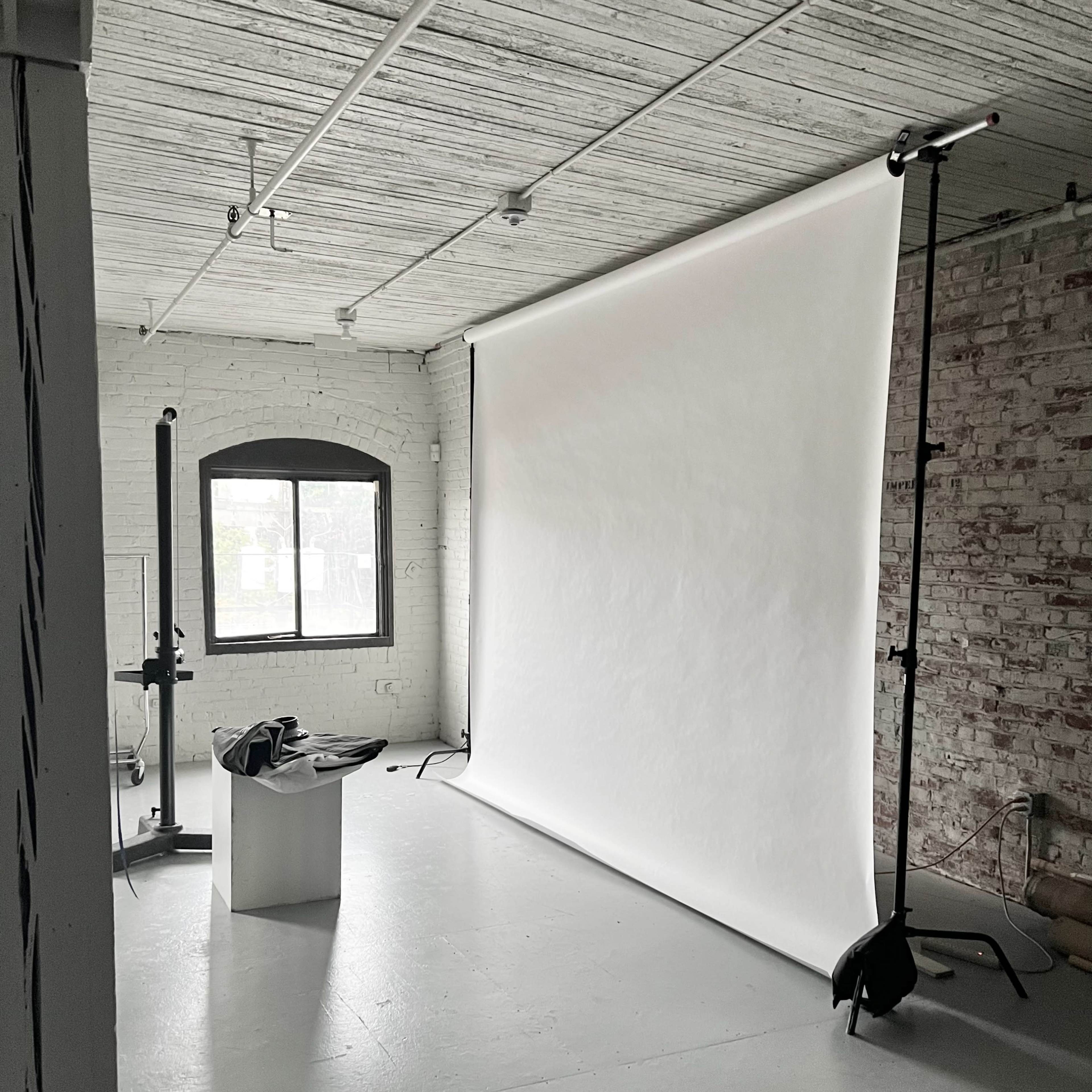 A photography studio features a large white backdrop, a stool, and minimal equipment set against a brick and plaster wall.