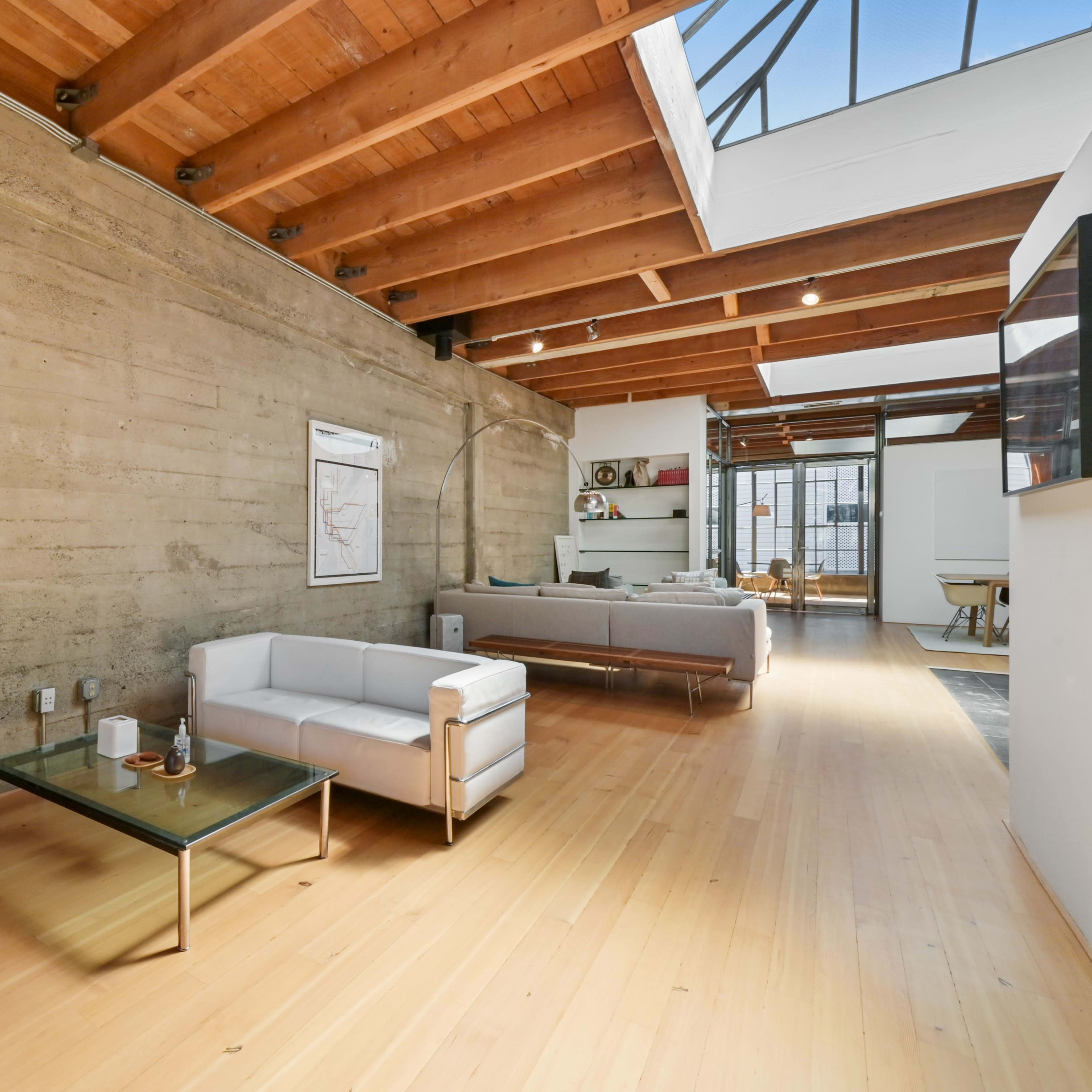 A spacious modern living area with concrete walls, wooden beams, and minimalist furniture arranged along a light hardwood floor.
