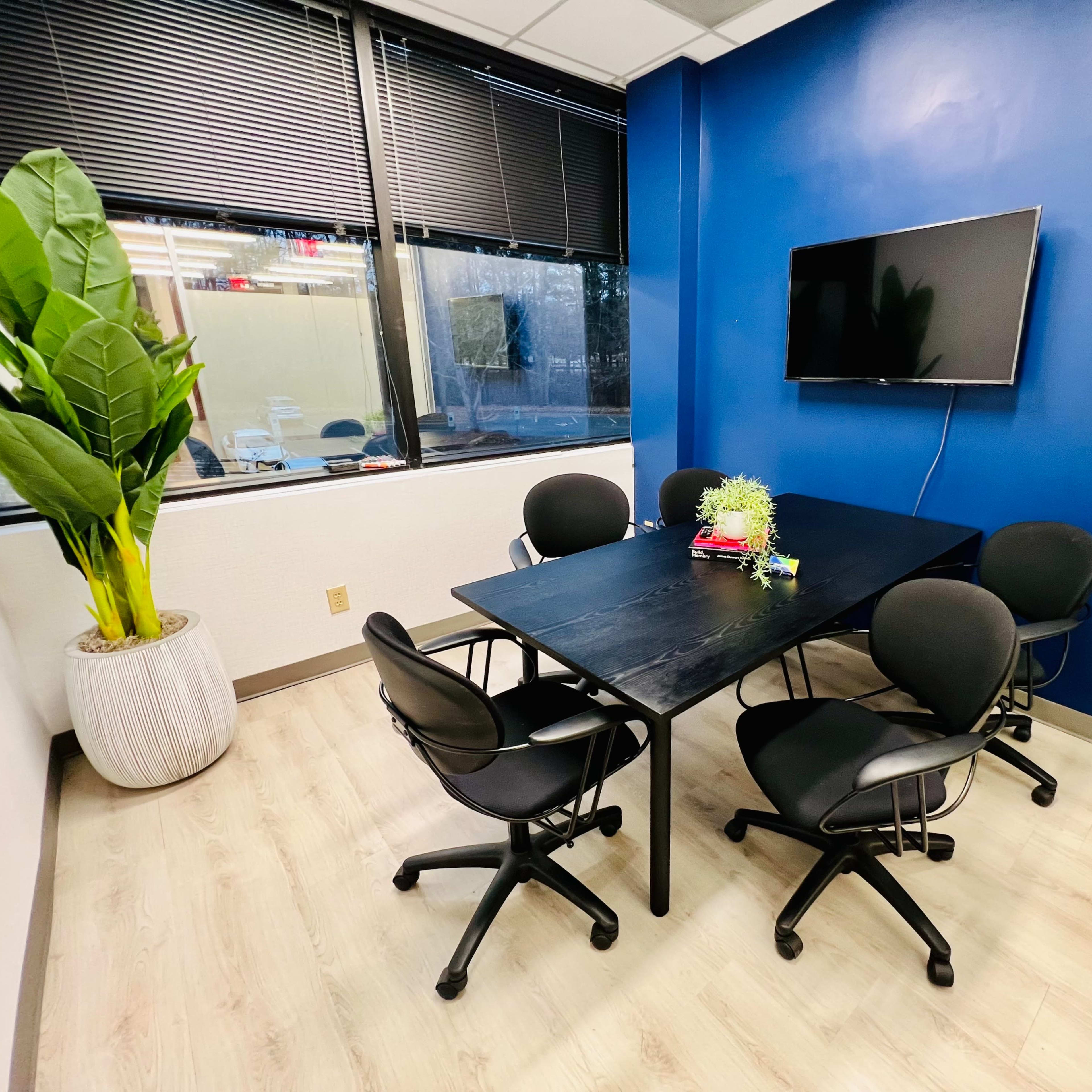 A small meeting room with a black table, five rolling chairs, a wall-mounted TV, a large plant in a pot, and blue walls.