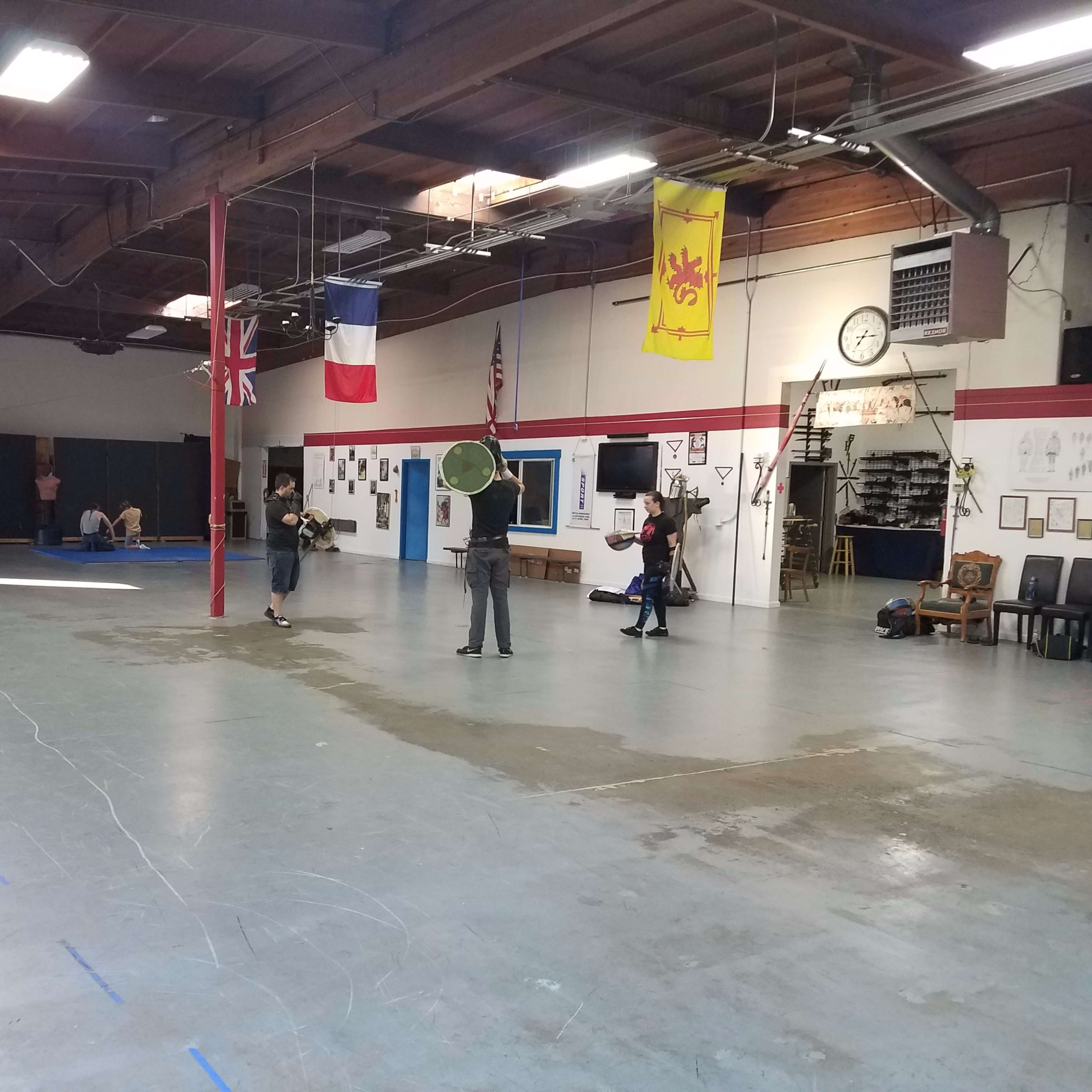 A group of individuals practices medieval combat techniques in a spacious indoor training area decorated with various flags.
