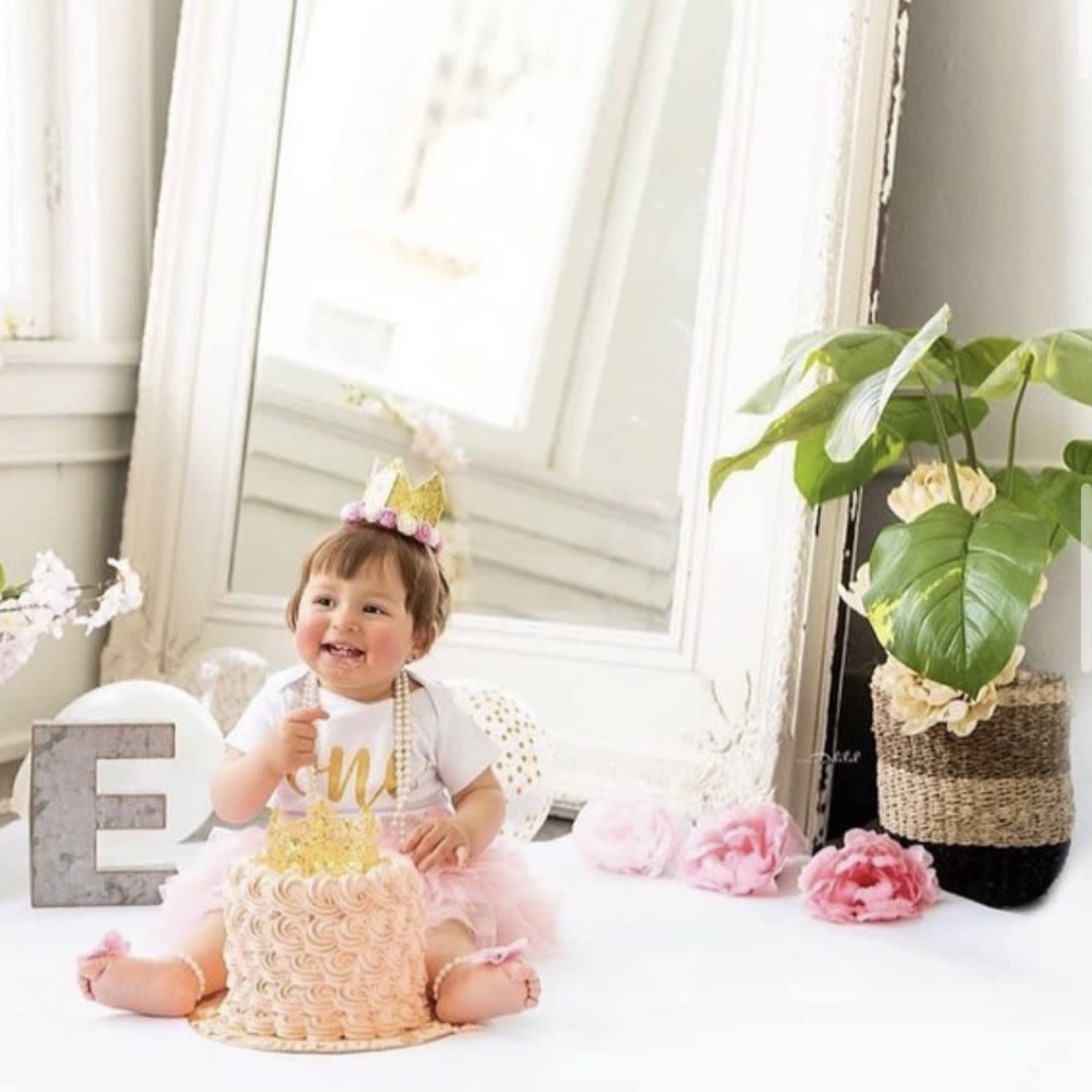 A child sits on a white surface next to a cake, surrounded by decorative flowers and the word "ONE" displayed in large letters.