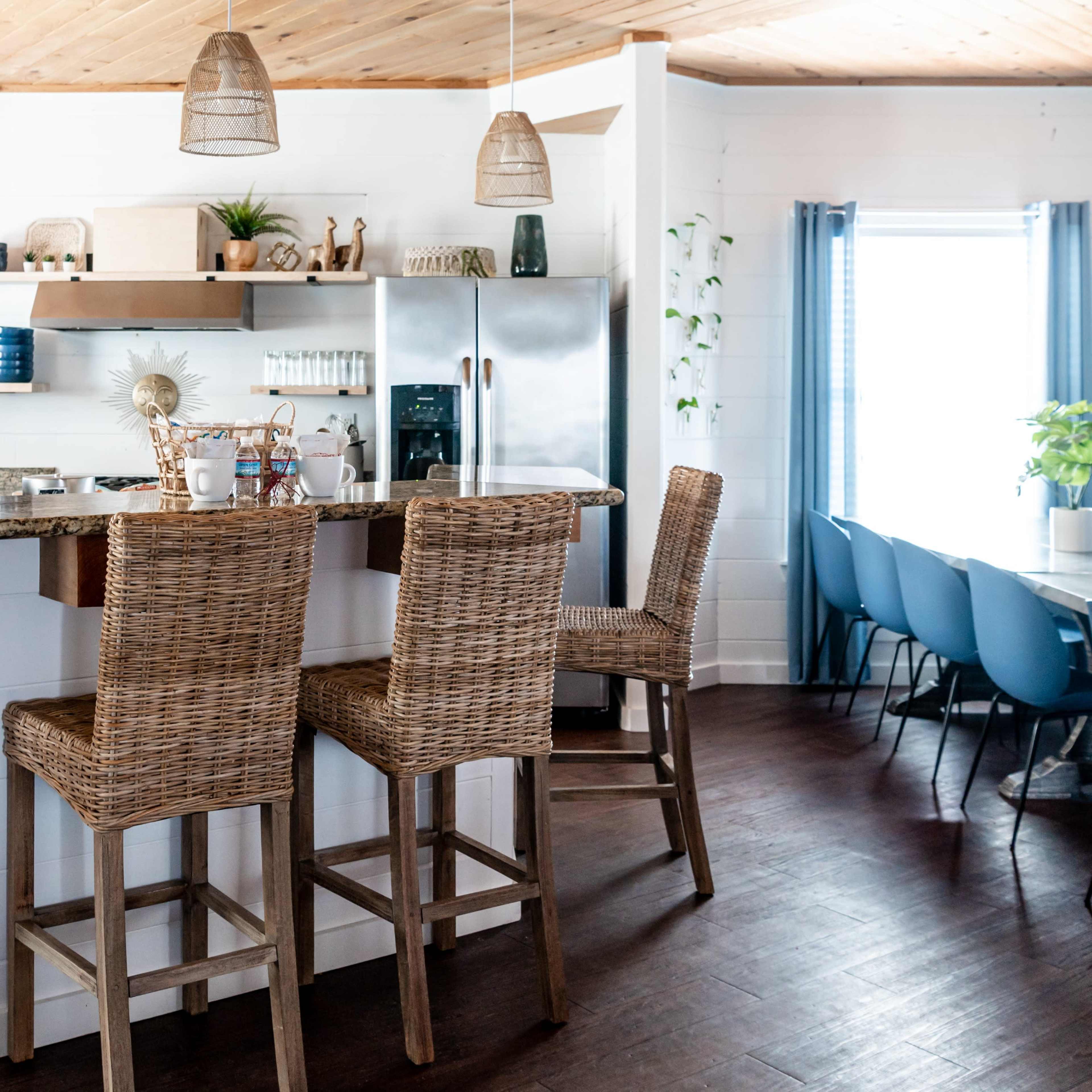 A modern kitchen features a bar with wicker stools and a dining table with blue chairs, complemented by light wood accents and plants.