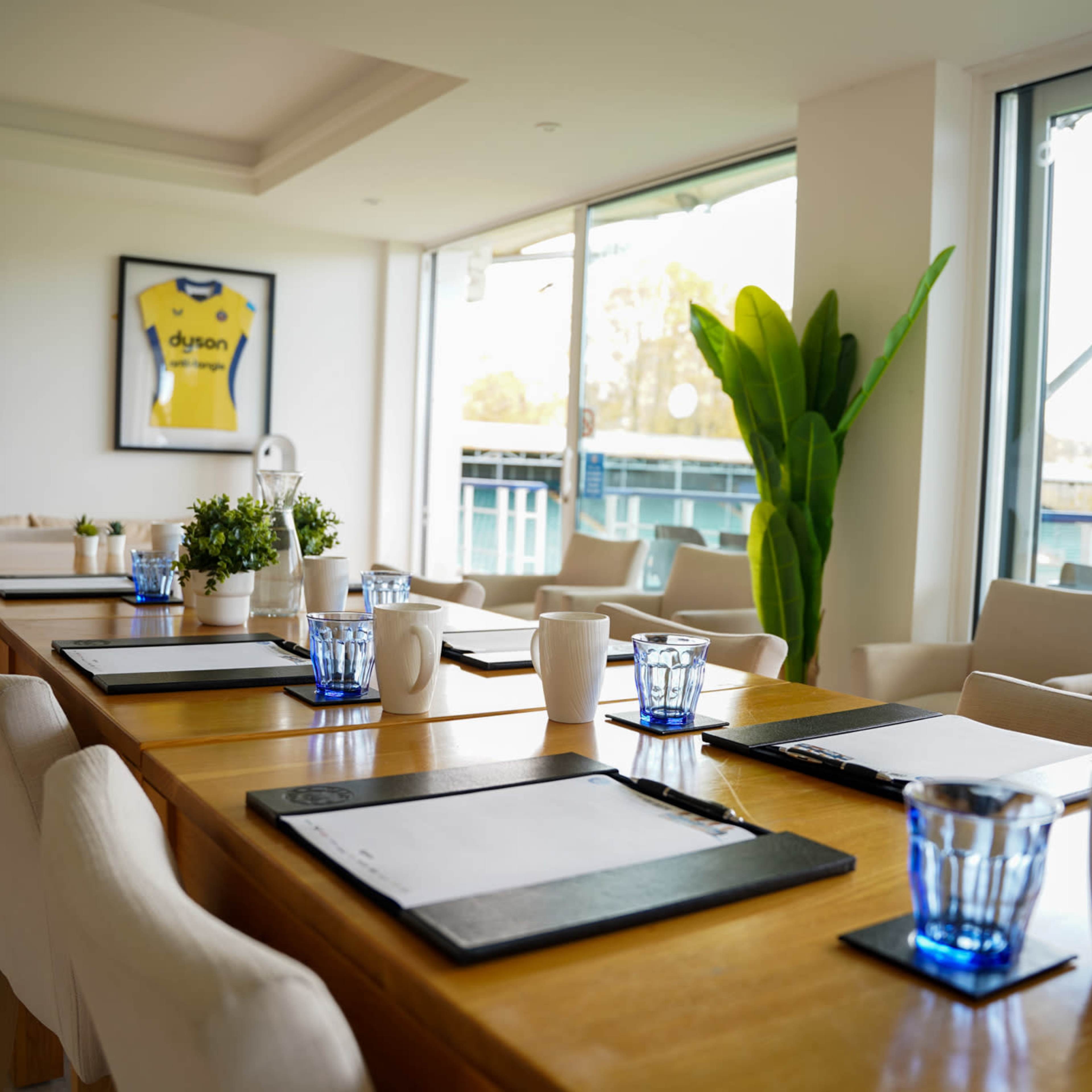 A modern meeting room features a long wooden table set with notepads and glasses, surrounded by comfortable chairs and framed jerseys on the walls.
