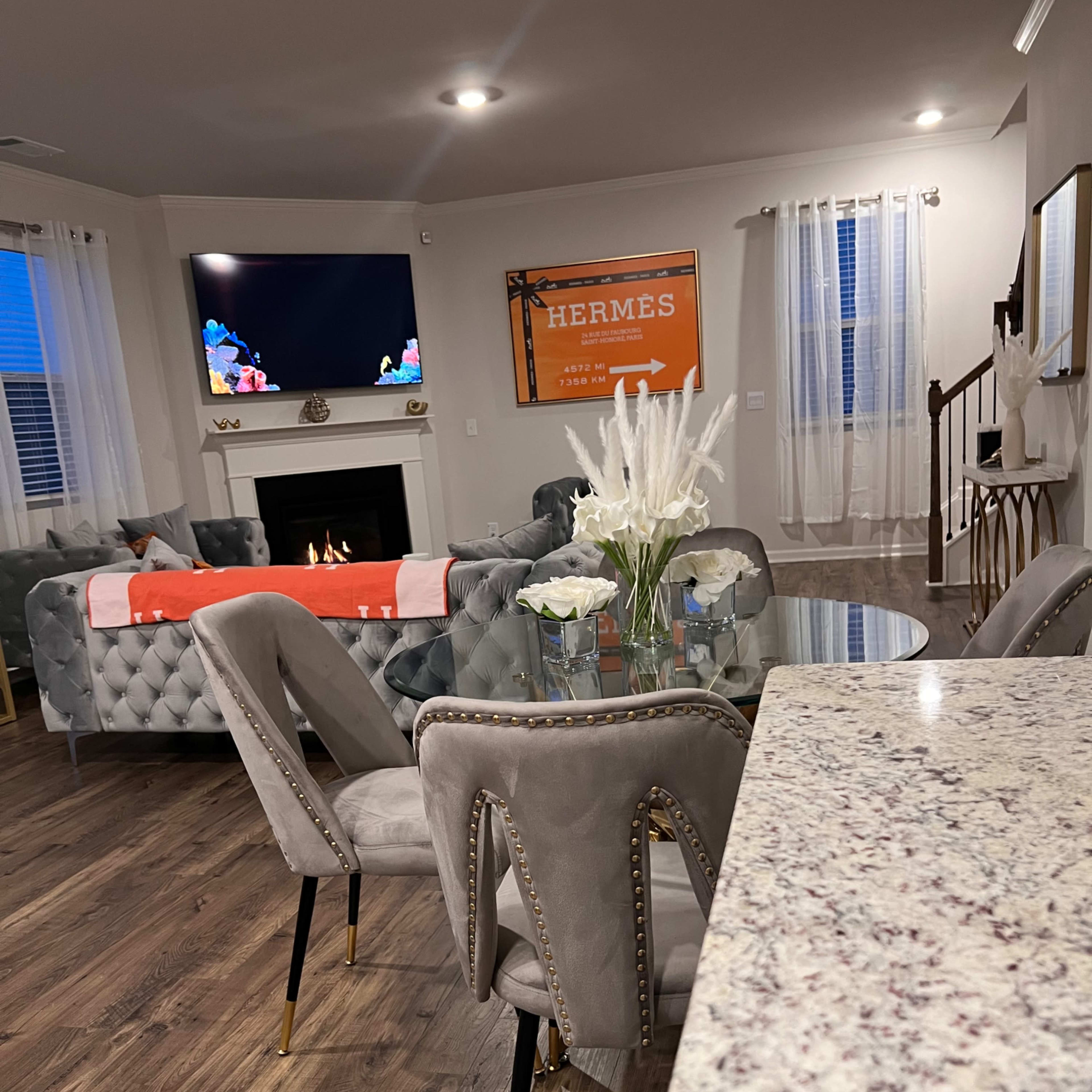 A modern living room features a glass dining table surrounded by upholstered chairs, with a TV mounted above a fireplace and orange wall art in the background.