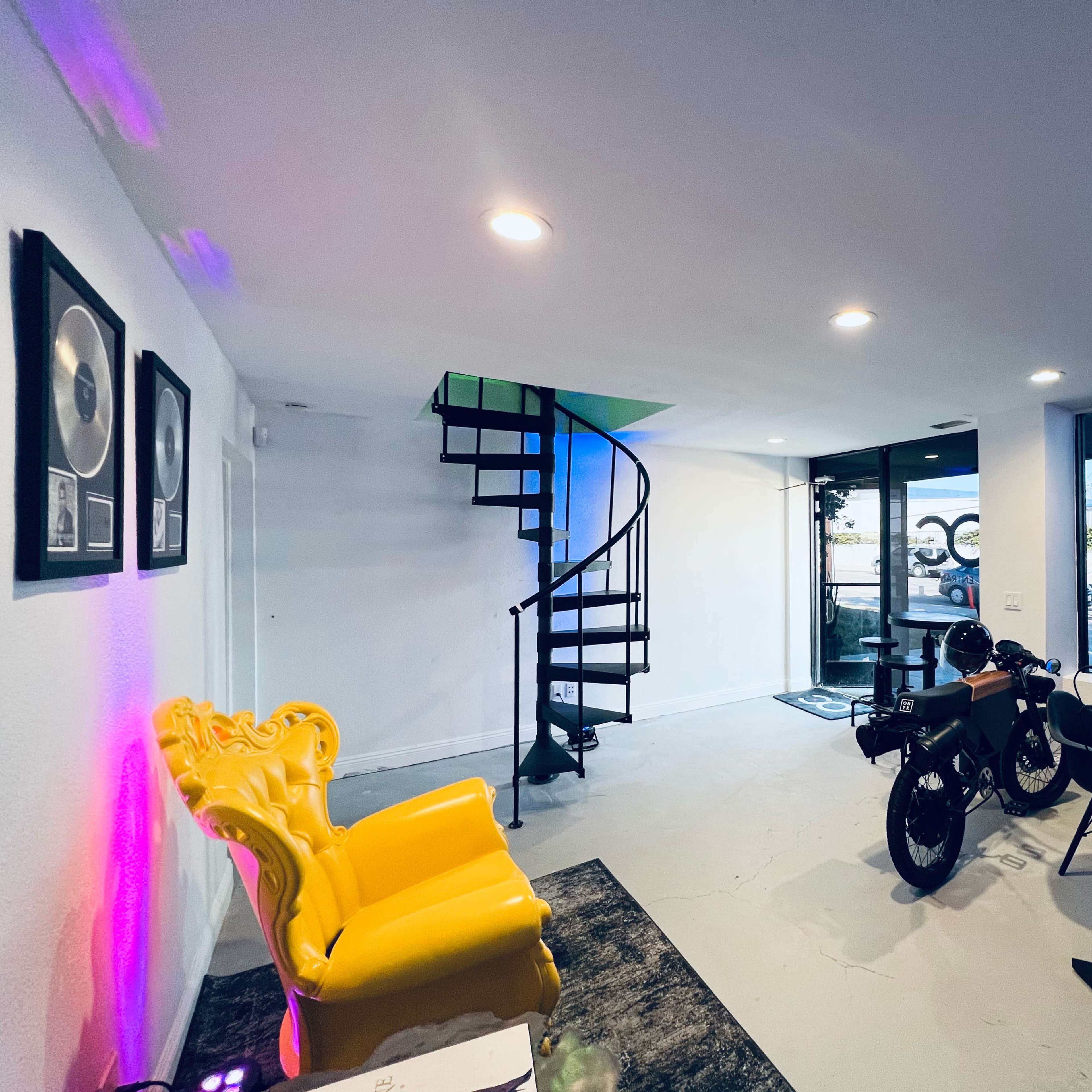 A bright, modern space features a yellow chair, a spiral staircase, and workstations near a large window.