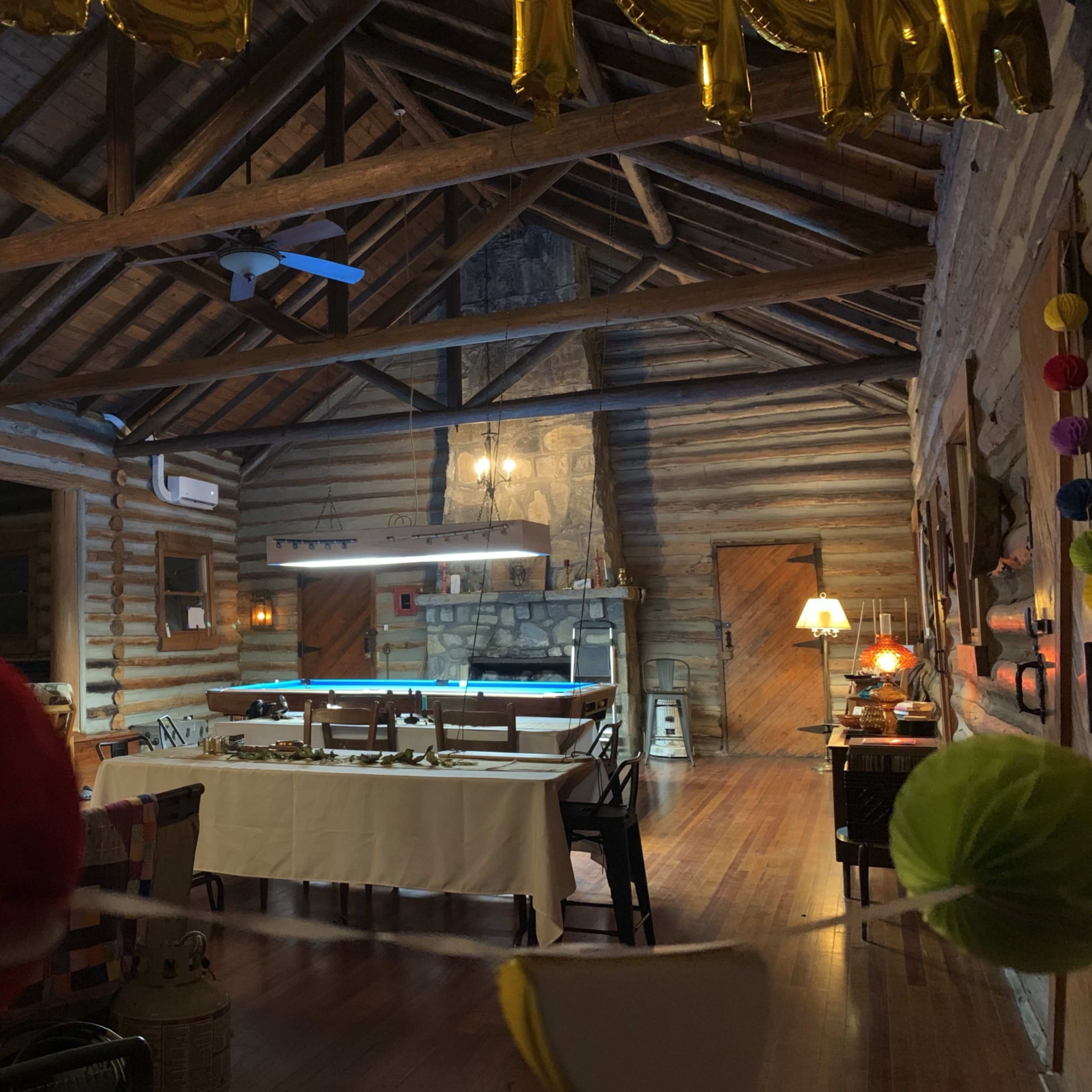 A spacious wooden interior of a rustic cabin, featuring exposed beams, a dining area with tables, and a stone fireplace.