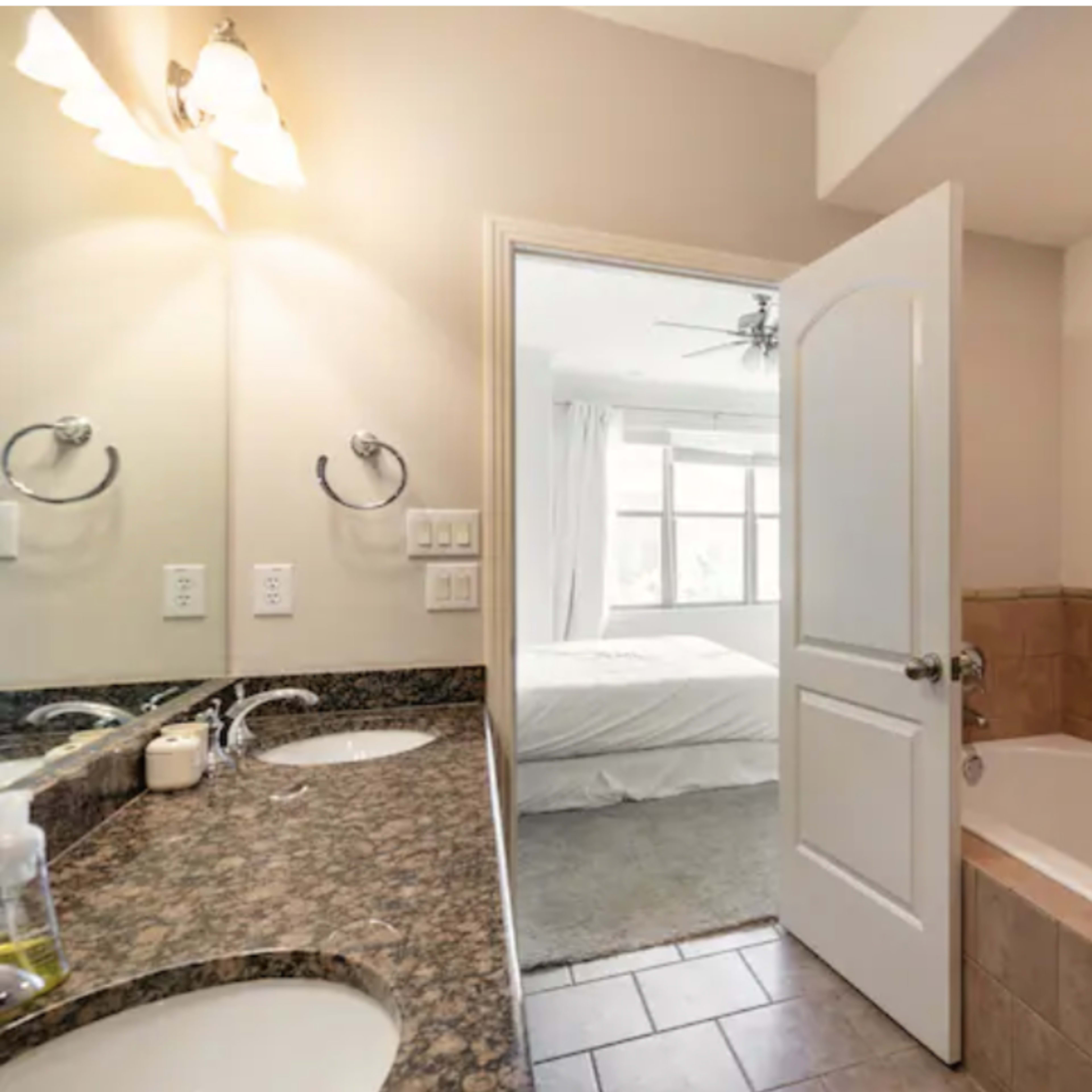 A bathroom featuring a double vanity with a mirror, a jetted tub, and an open door leading to a bedroom with a bed and large window.
