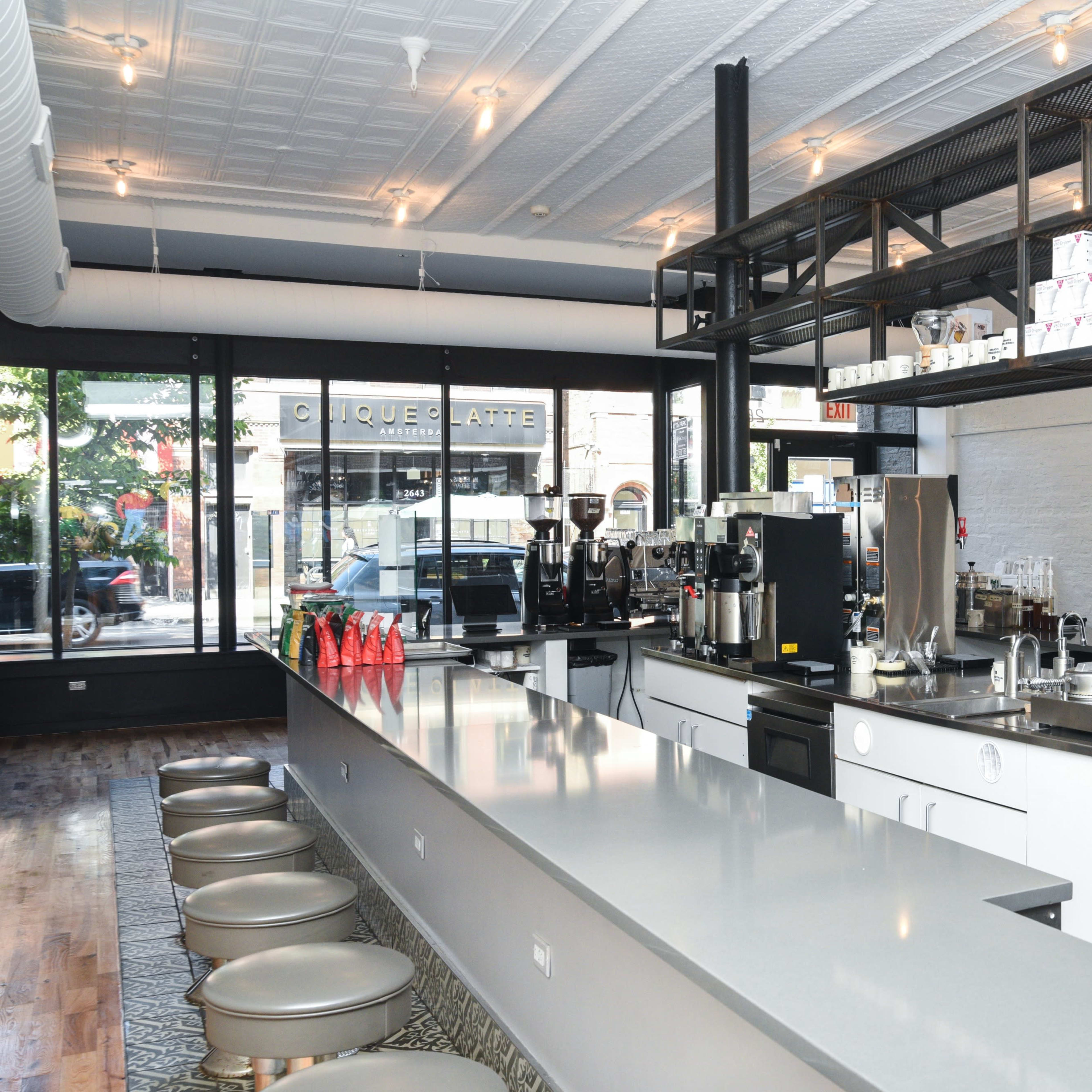 A modern, minimalist café interior featuring a long counter with coffee machines and seating in front of large windows.