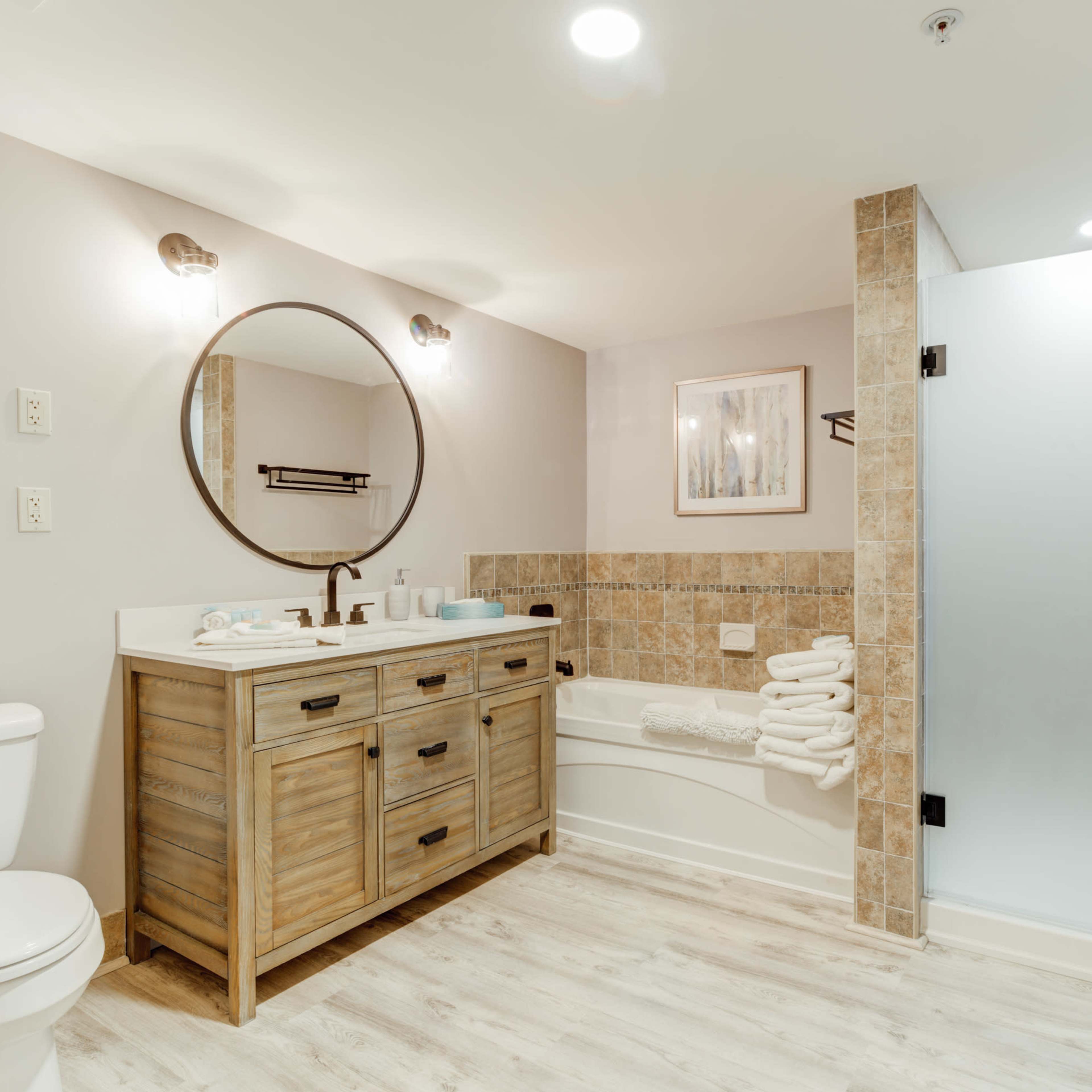 The bathroom features a wooden vanity with a round mirror, a bathtub with beige tiles, a glass shower enclosure, and a toilet.