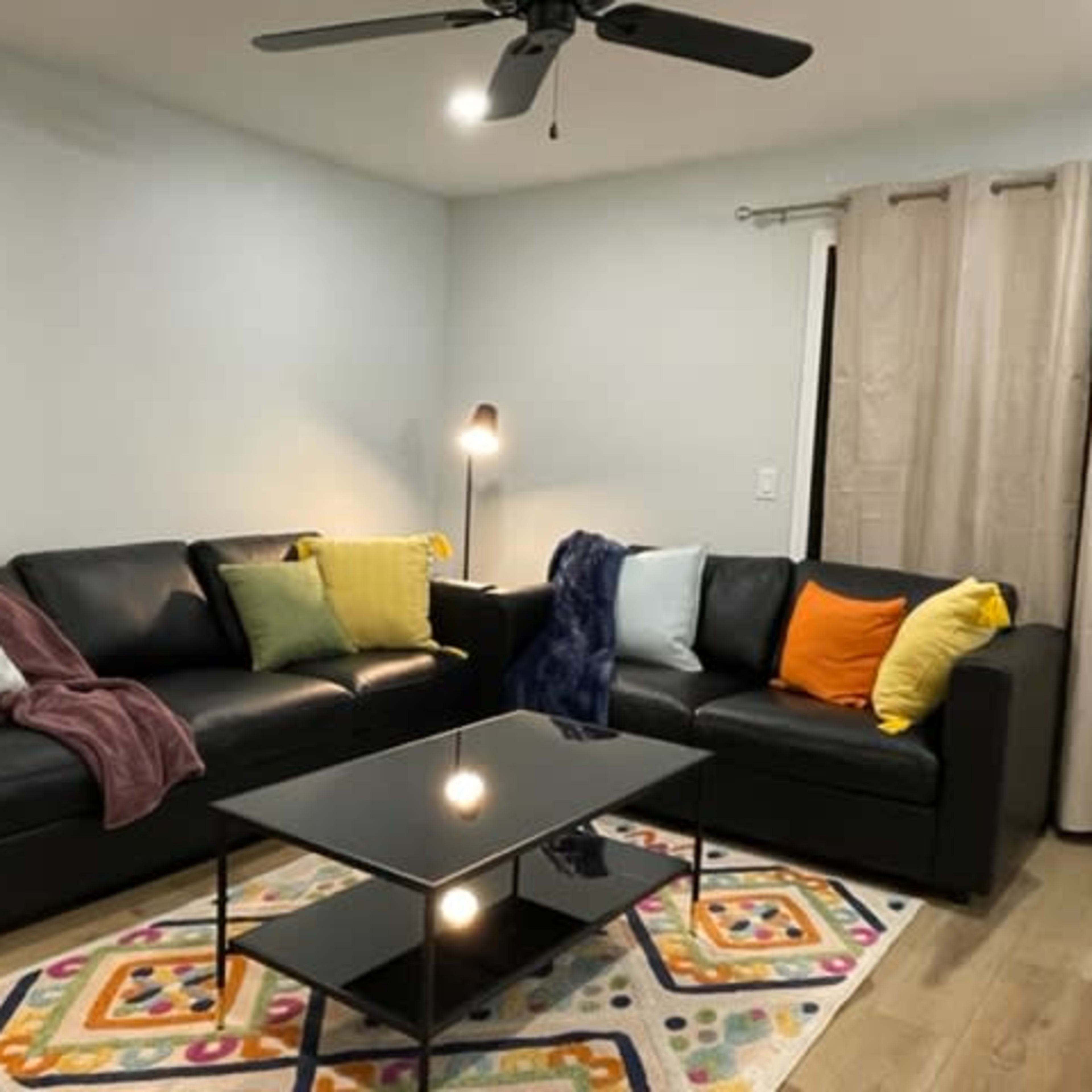 The room features a black leather sectional sofa adorned with colorful pillows and a coffee table placed on a patterned area rug.