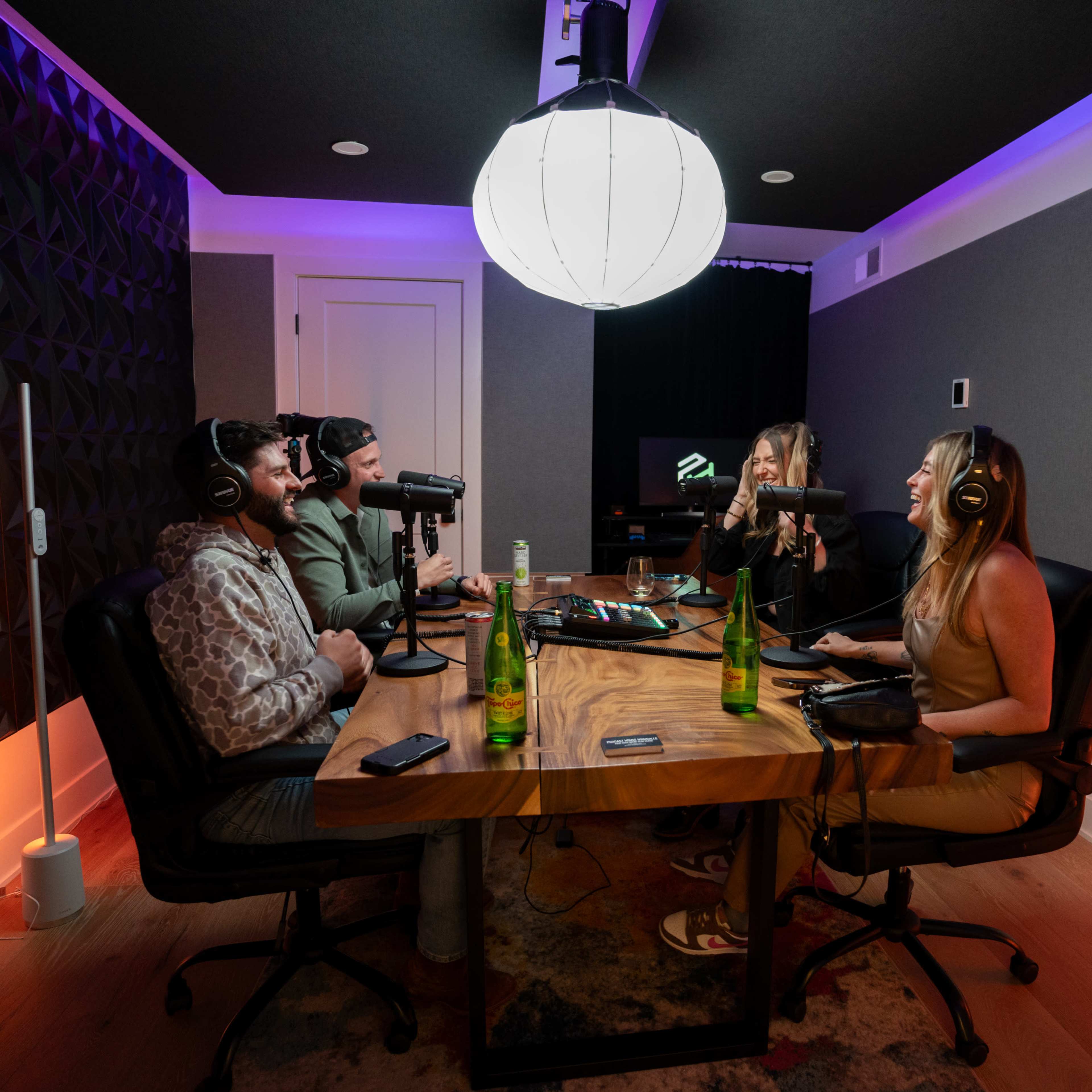 Four people sit around a table in a podcast studio, equipped with microphones and drinks, engaged in conversation under a large overhead light.