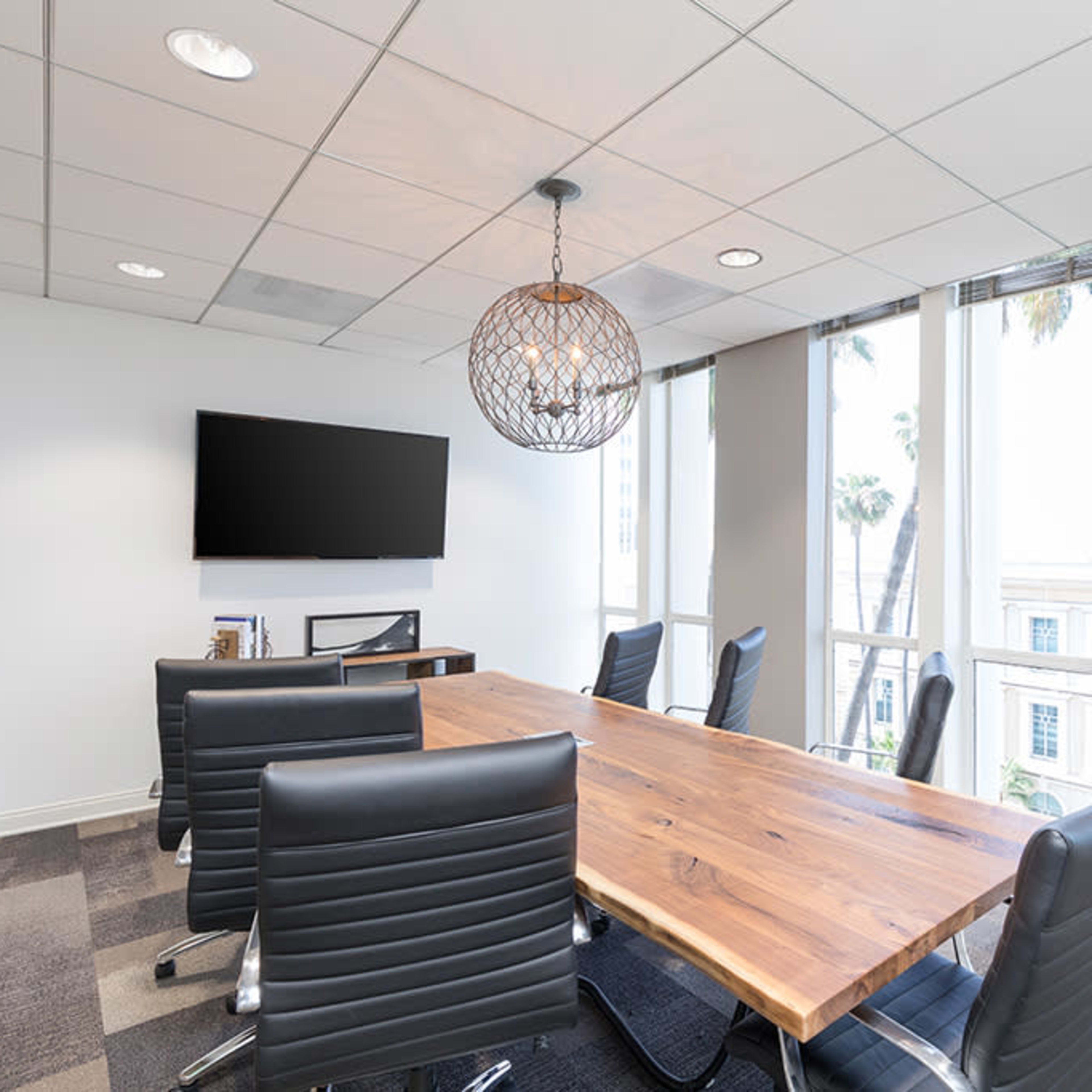 A modern conference room featuring a large wooden table, black leather chairs, and a wall-mounted television beside large windows.