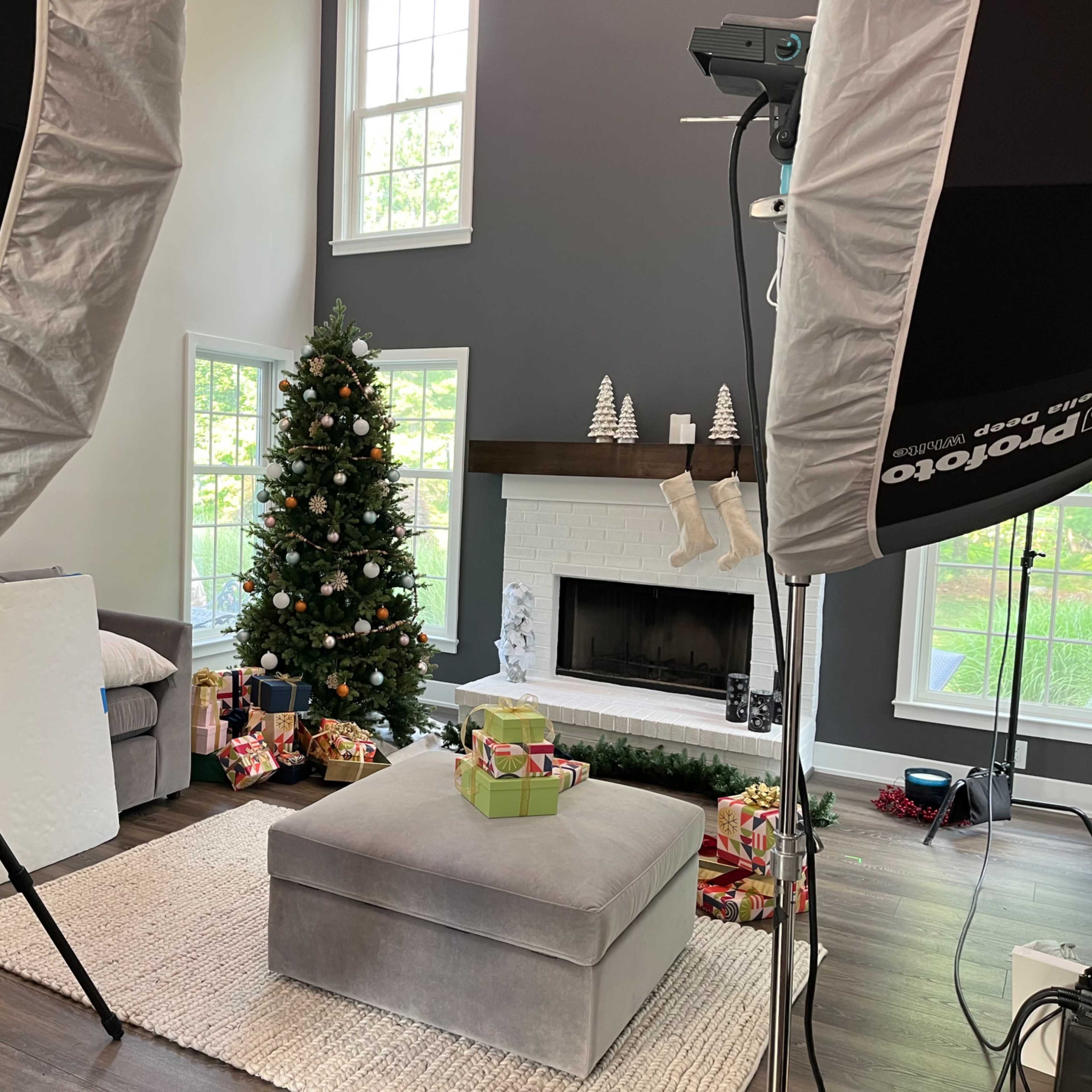 A living room decorated for the holidays, featuring a Christmas tree, a coffee table with presents, and large softbox lights set up for a photo shoot.