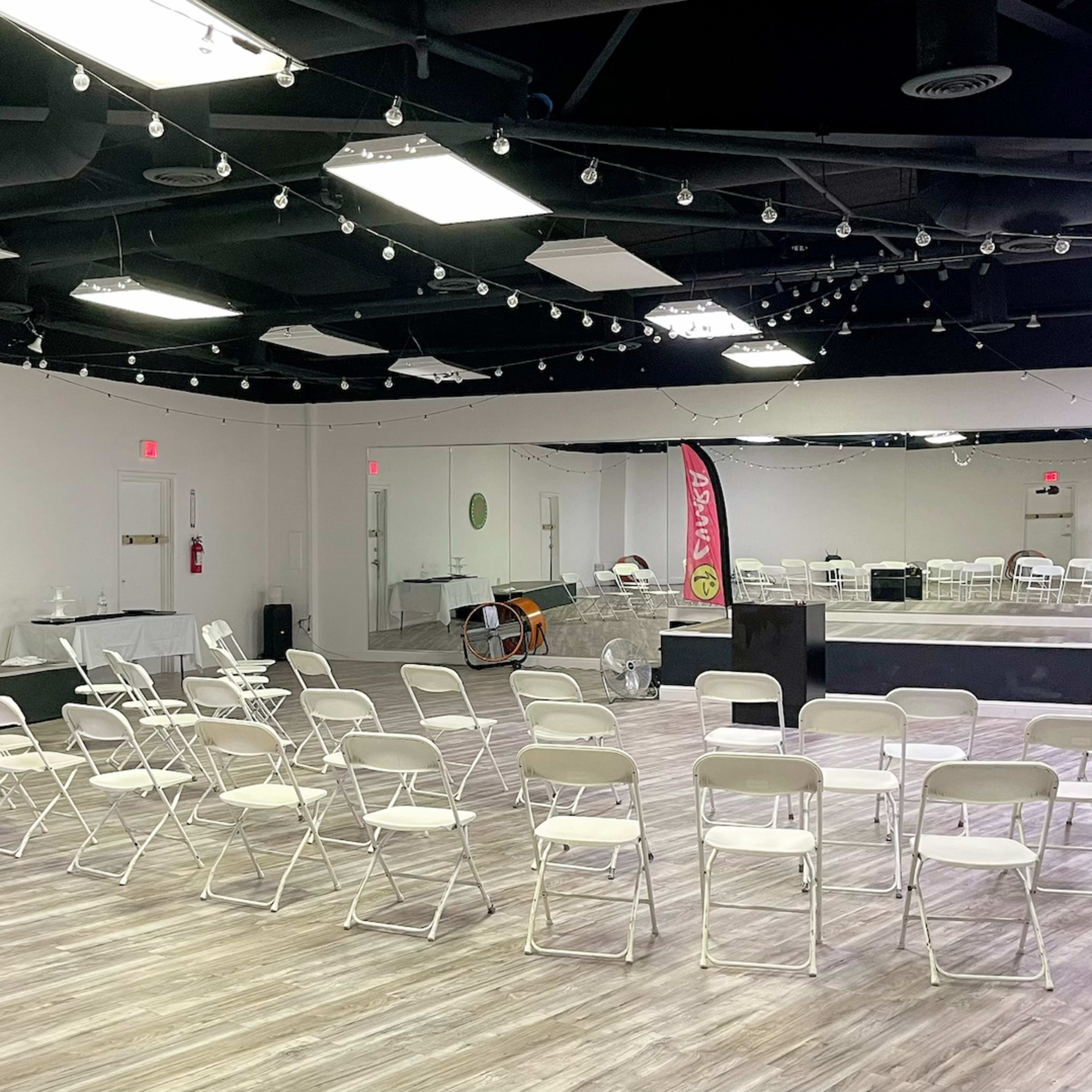 A spacious, well-lit event room with rows of white chairs facing a stage area, complemented by mirrors and decorative elements on the walls.