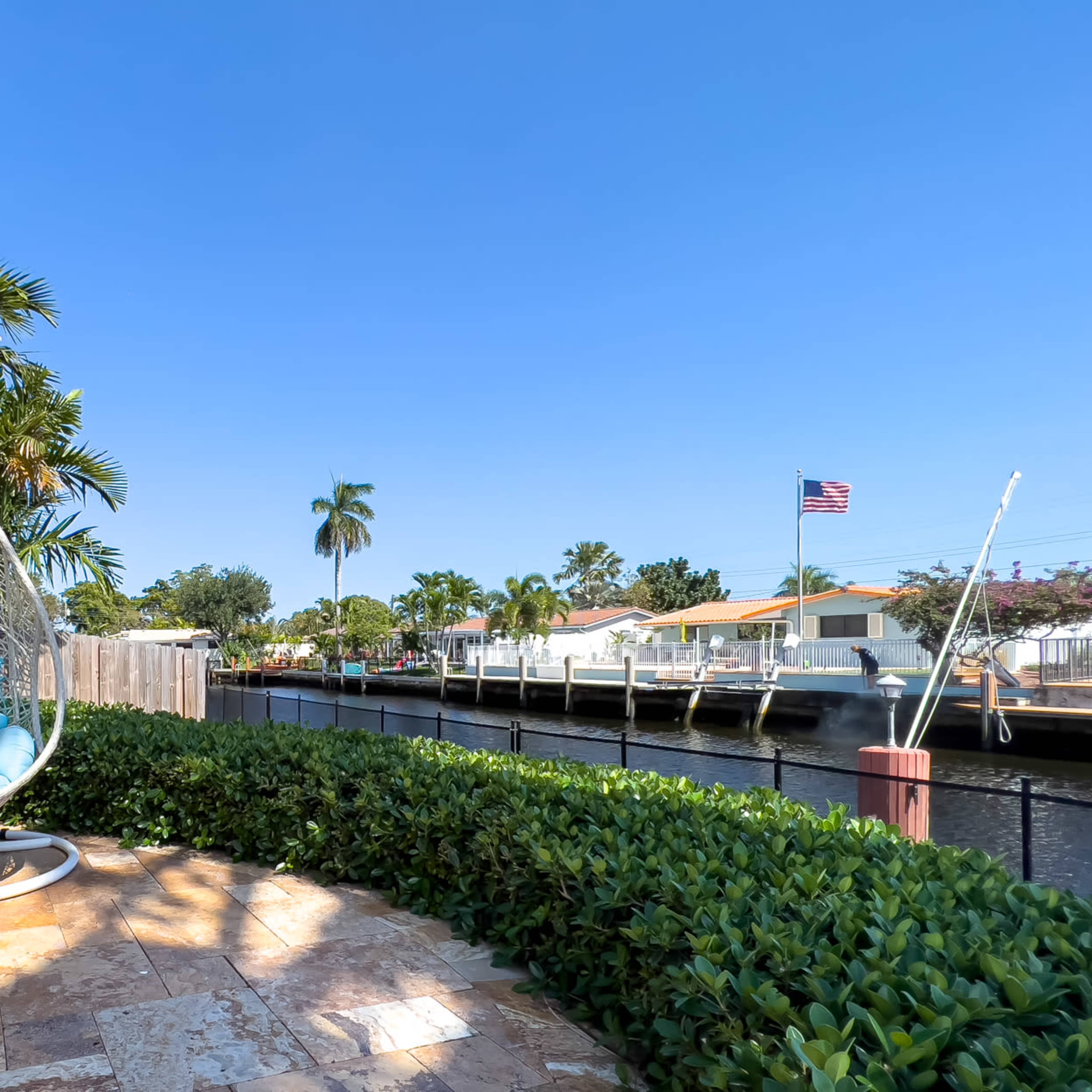 A hanging chair with blue cushions is positioned on a patio overlooking a canal lined with palm trees and houses.