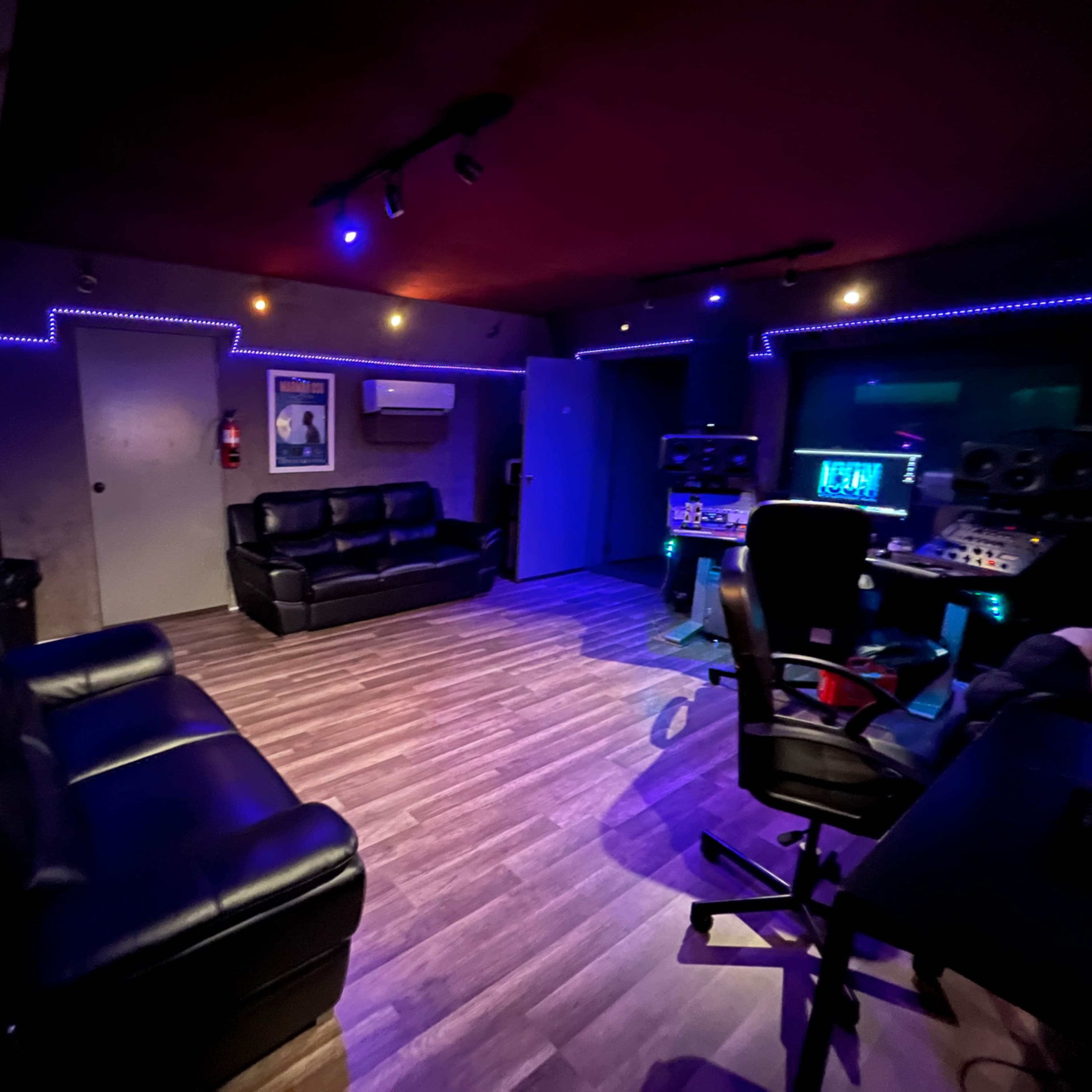 A dimly lit music studio featuring black leather sofas, a desk with studio equipment, and ambient lighting along the walls.