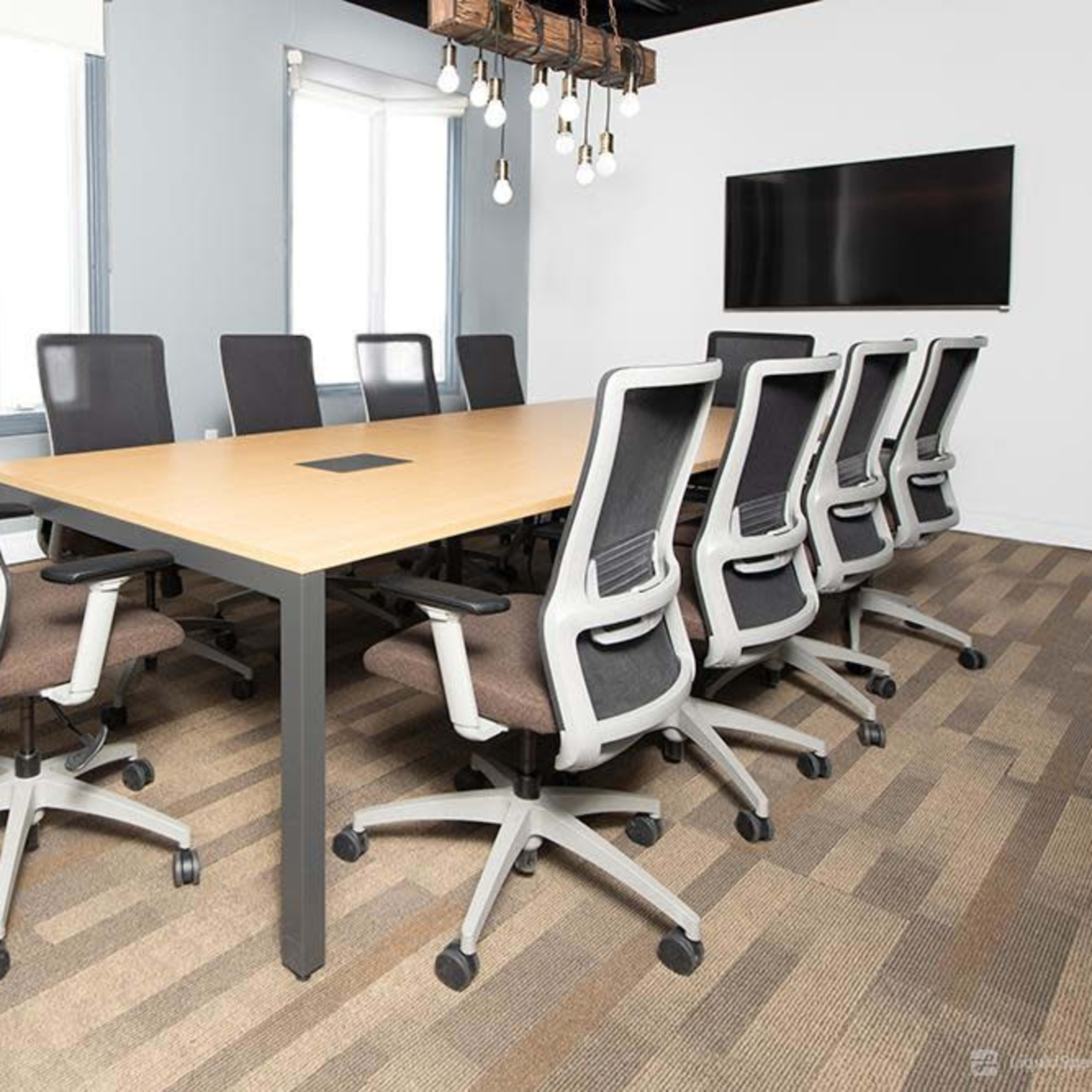 A large conference room features a rectangular wooden table surrounded by ergonomic office chairs, with windows and a wall-mounted screen visible in the background.