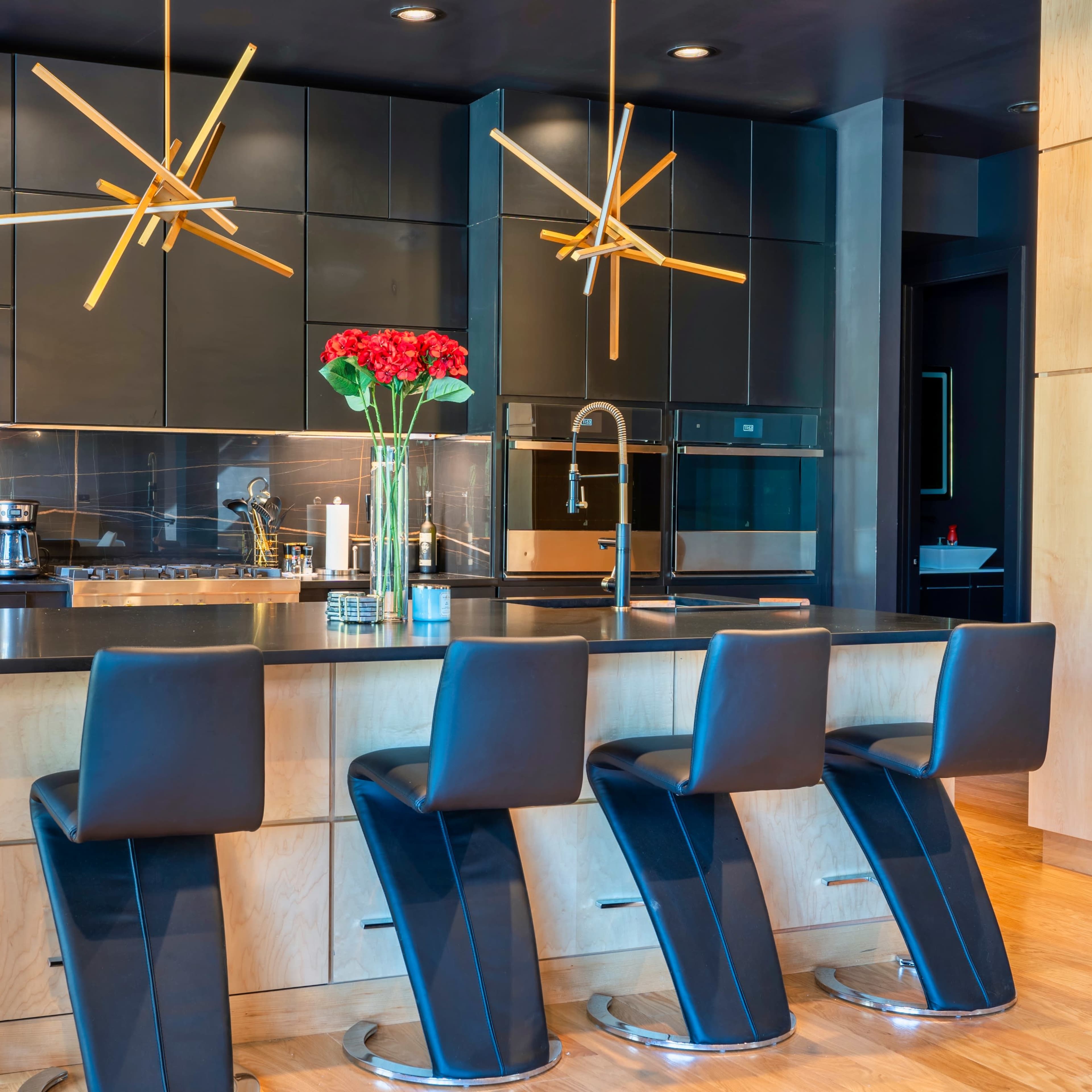A modern kitchen features a central island with four black stools, sleek cabinetry, and decorative light fixtures overhead.