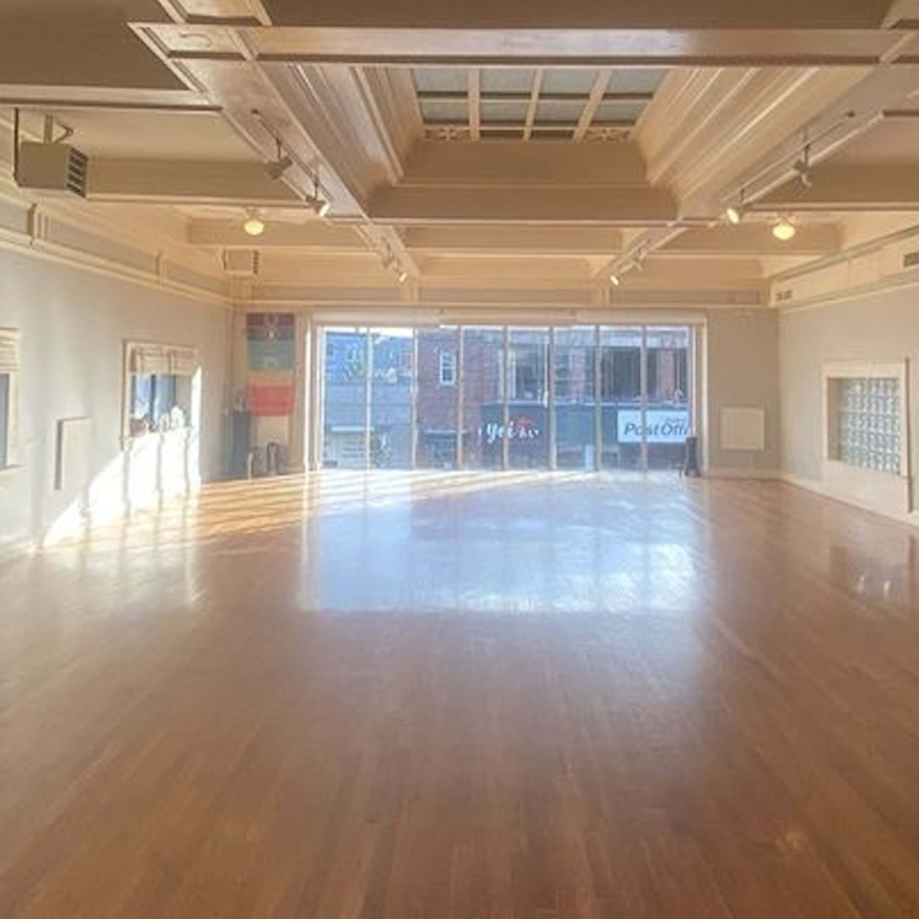 A spacious, sunlit studio with wooden floors and large windows overlooking an urban street.